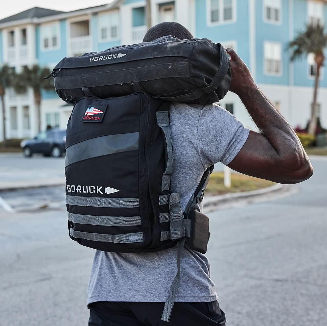 Man carrying black GORUCK rucking backpack and sandbag outdoors near blue buildings