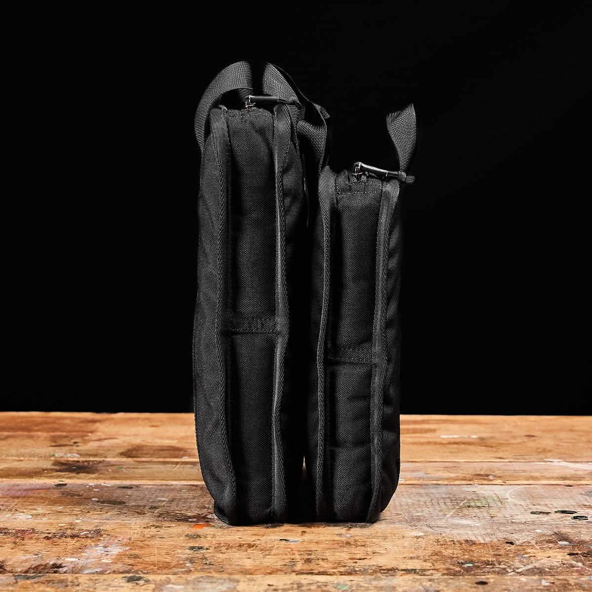 Two black fabric cases, one slightly taller and wider than the other, stand upright on a rustic wooden surface against a black background. Their sturdy design mirrors the resilience of Sand Ruck Plates by GORUCK, providing sleek functionality and the assurance of enduring quality.