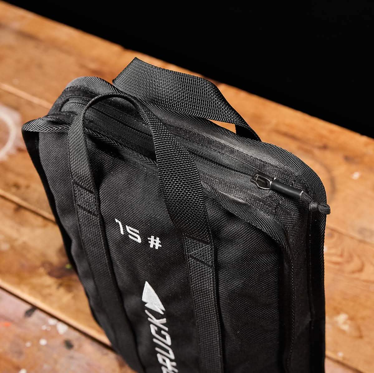 A black backpack with zippers and handles, adorned with the numeral 15 and a logo similar to RUCK, is tailored for adventurers seeking to carry their Sand Ruck Plates. Displayed on a rustic wooden surface, this GORUCK product comes with a lifetime guarantee for assurance.