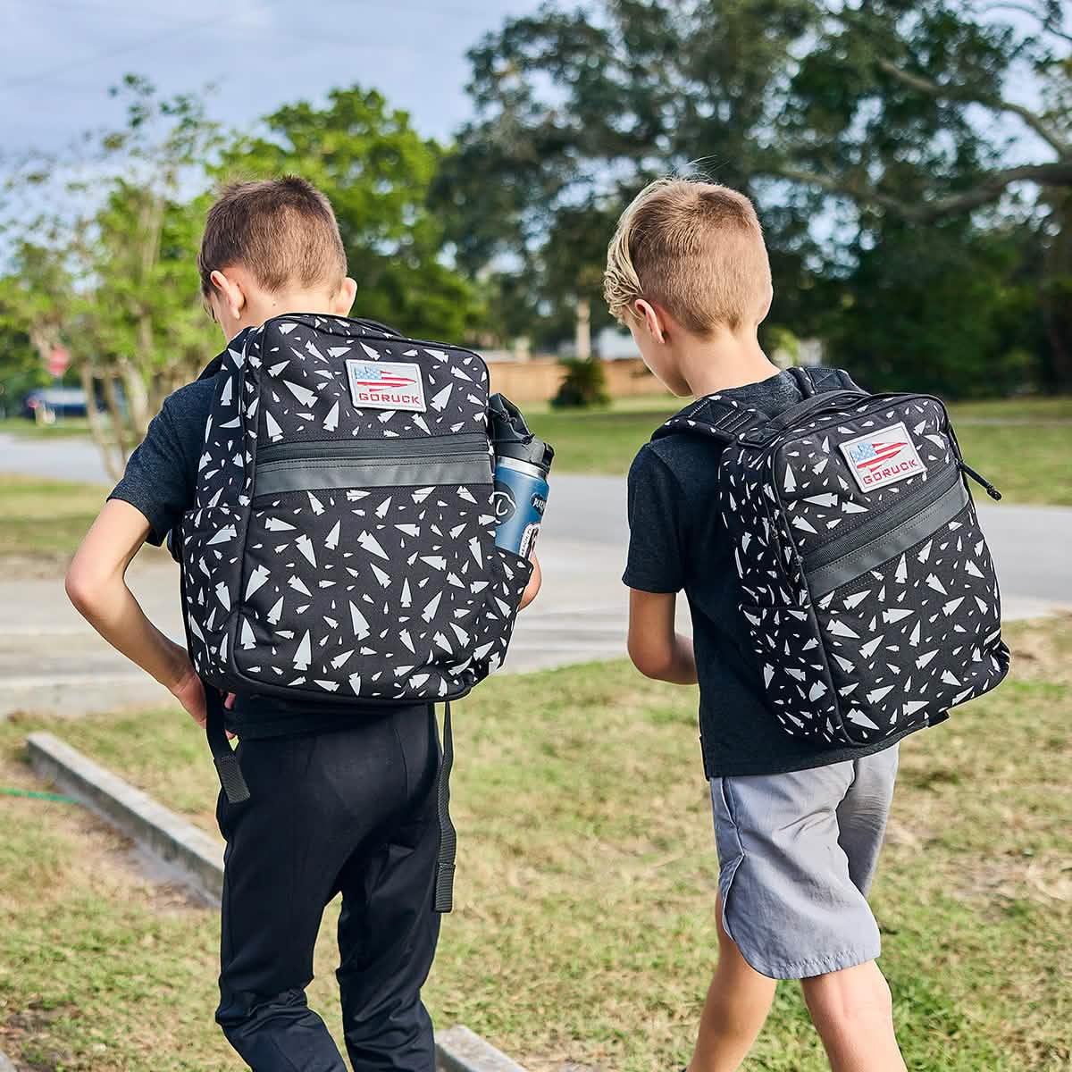 Two boys wearing matching REFLECTIVE KR1 black-and-white patterned school backpacks walk outside on a sunny day.