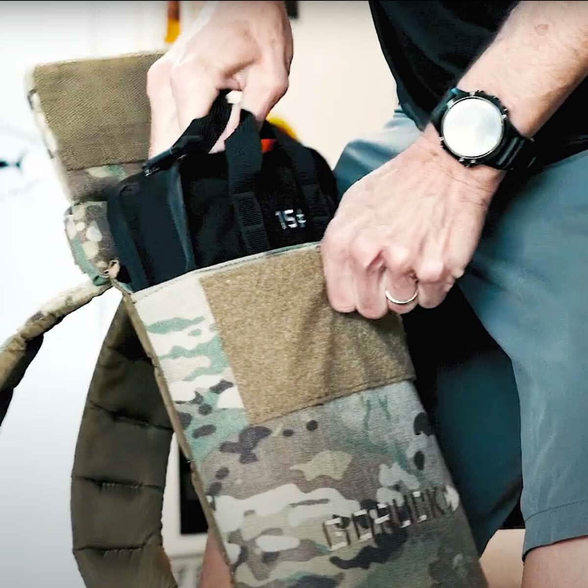 A person with a watch is putting a black item, likely a Sand Ruck Plate from GORUCK, into a camouflage pouch featuring shoulder straps. This tactical bag, improved with Velcro closures and durable straps, embodies the durability often associated with Lifetime Guarantee products.