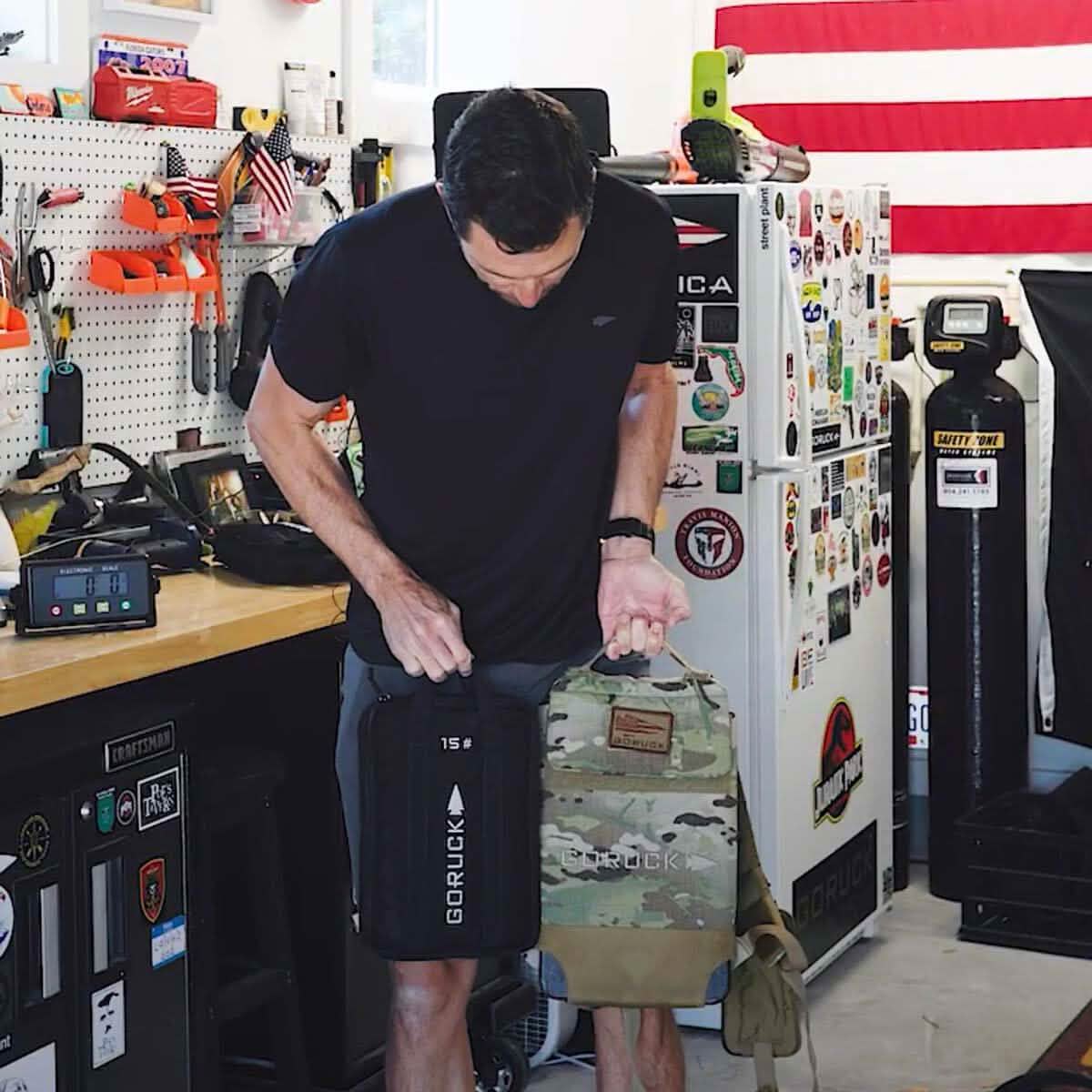 A person in a black t-shirt stands in a garage workshop holding two GORUCK backpacks, each designed to accommodate Sand Ruck Plates. The room is adorned with an American flag and various stickers on a fridge, with tools and equipment visible on the workbench and walls—a testament to durability deserving of a lifetime guarantee.