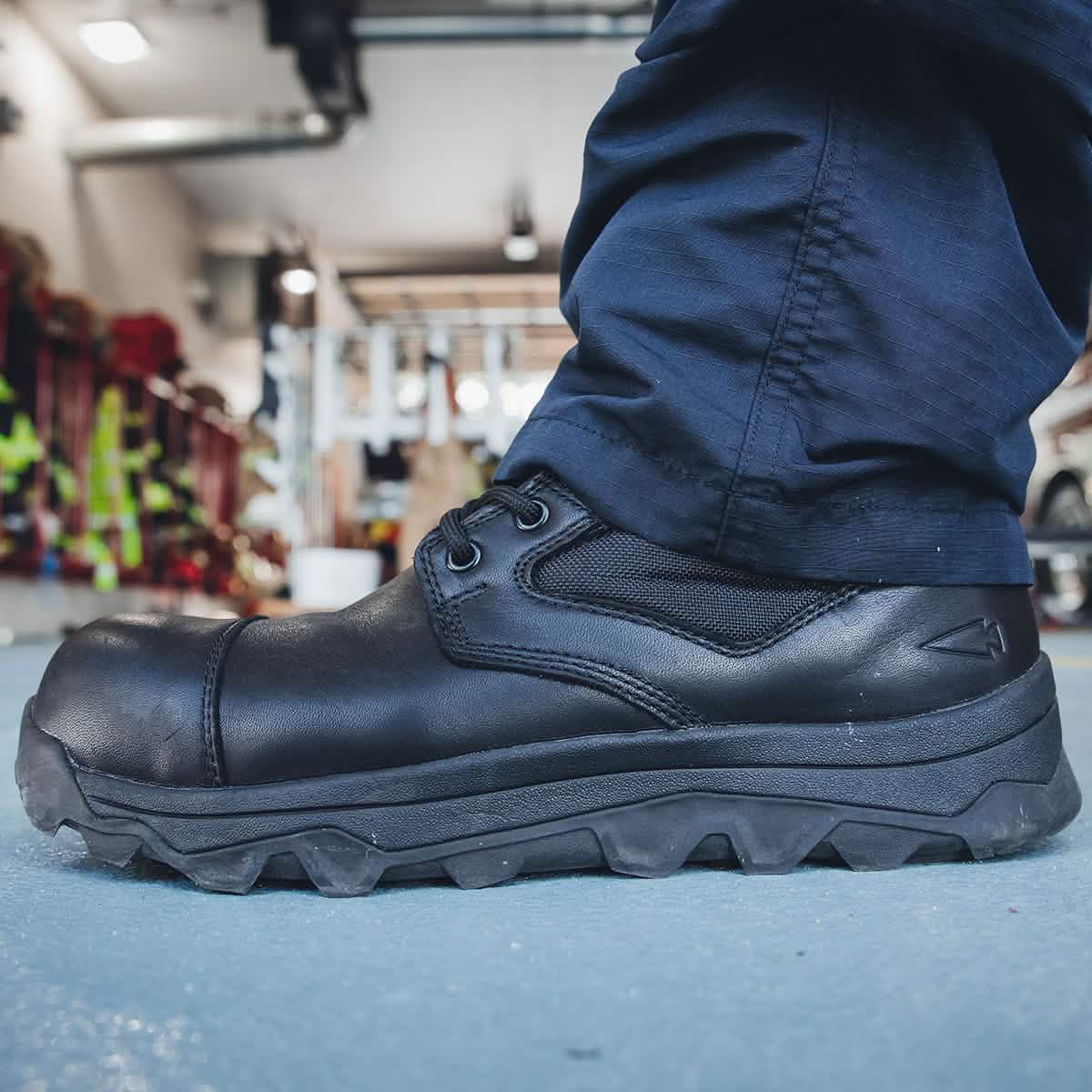 A person wearing the MACV-2 Safety Boot - High Top by GORUCK stands on a blue-gray floor in a garage or workshop setting. Crafted to meet Special Forces standards, gear and equipment can be seen in the blurry background.