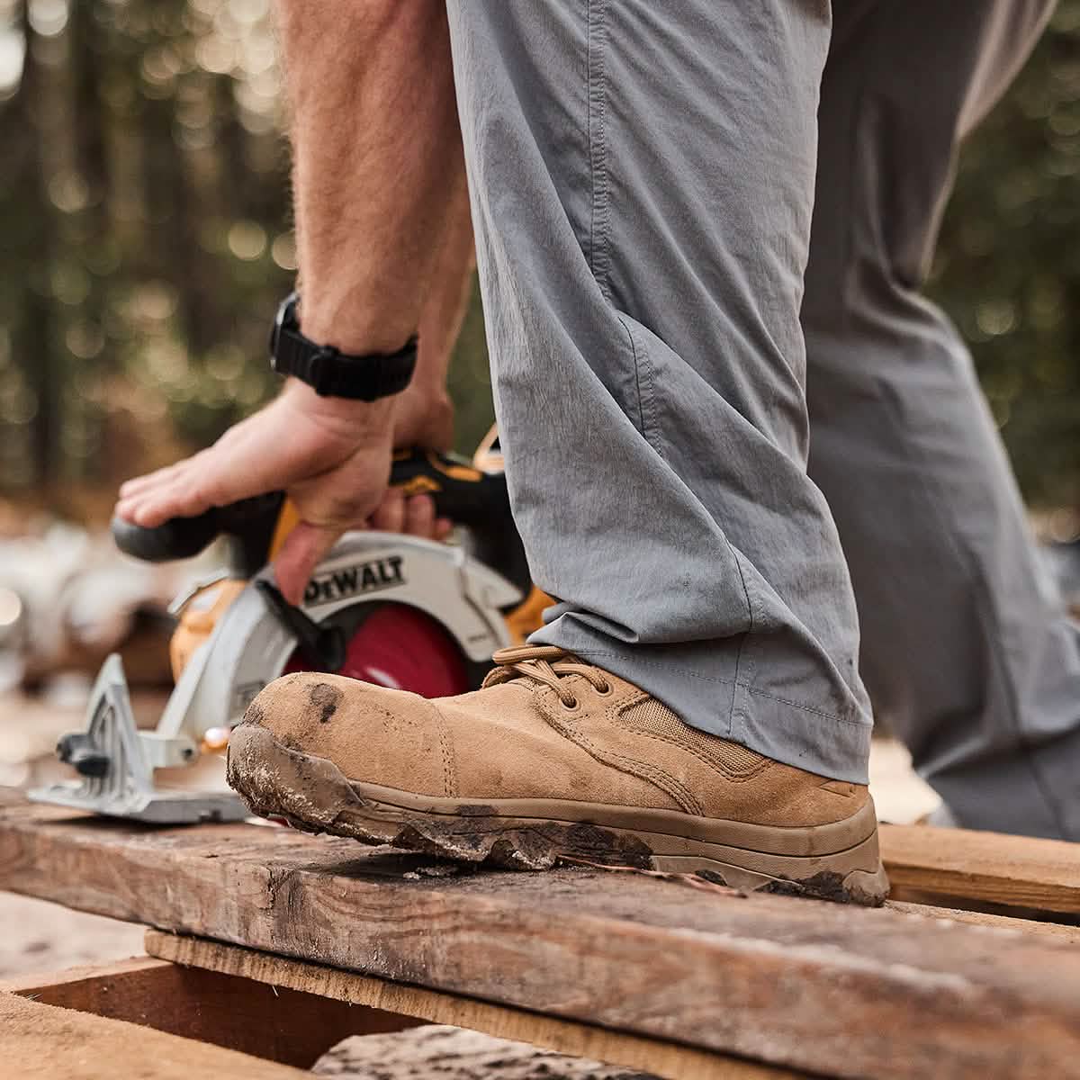 Wearing GORUCK's MACV-2 Safety Boot - Mid Top, a person clad in gray pants carefully maneuvers a circular saw over wooden planks. The forest backdrop with blurred trees hints at outdoor construction or woodworking that demands the durability and skill worthy of Special Forces standards.