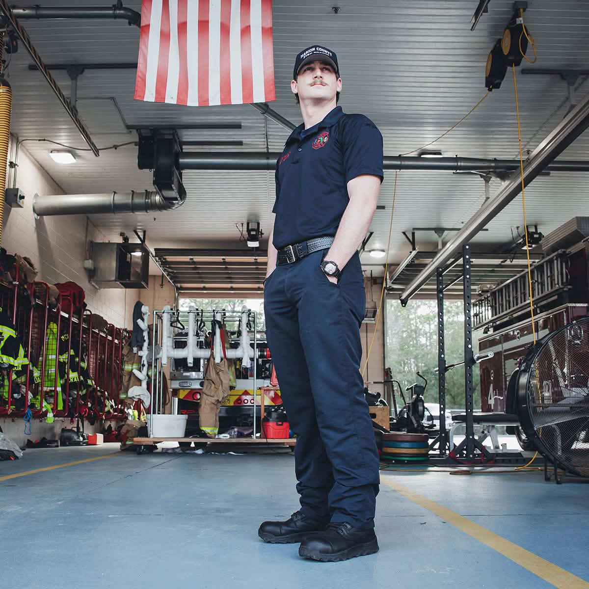 A firefighter in uniform, equipped with GORUCK's MACV-2 Safety Boot - High Top, stands confidently in a brightly lit fire station. An American flag hangs above, while firefighting equipment is visible in the background.