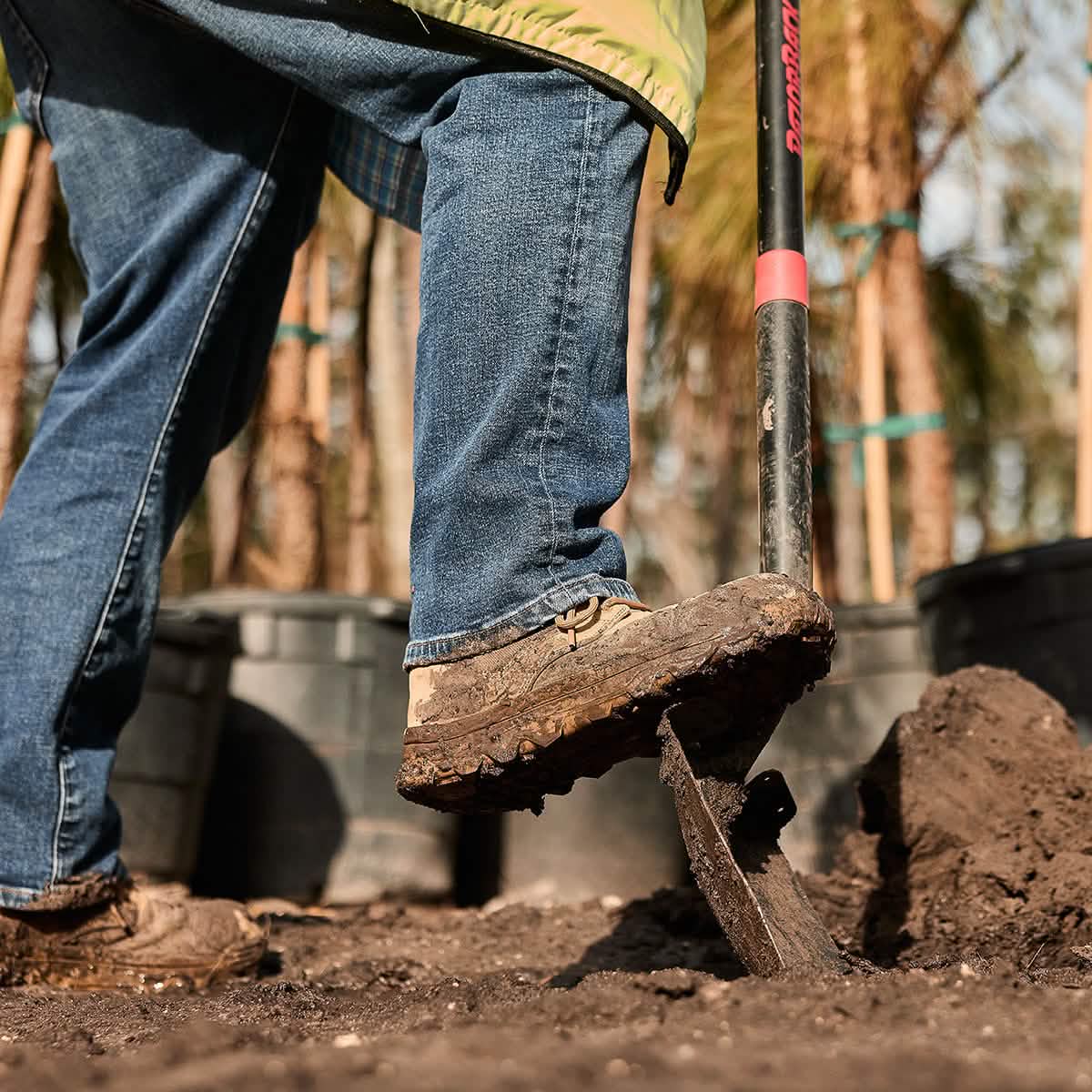 A person wearing jeans and the GORUCK MACV-2 Safety Boot - Mid Top uses a shovel to dig into the soil. Large potted plants and trees are visible in the background, suggesting a gardening or landscaping activity that meets Special Forces standards of precision and care.