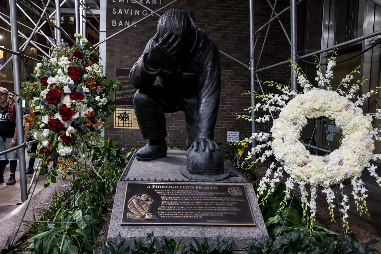 A firefighter memorial statue kneels with head in hand, surrounded by flower wreaths and scaffolding.