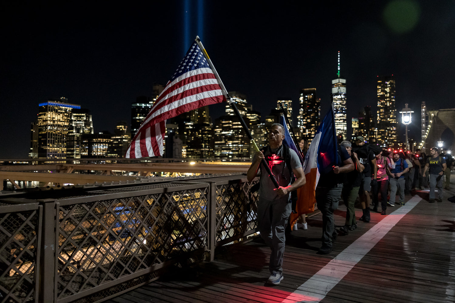 People walk across a bridge at night, carrying American and state flags, with city lights in the background.