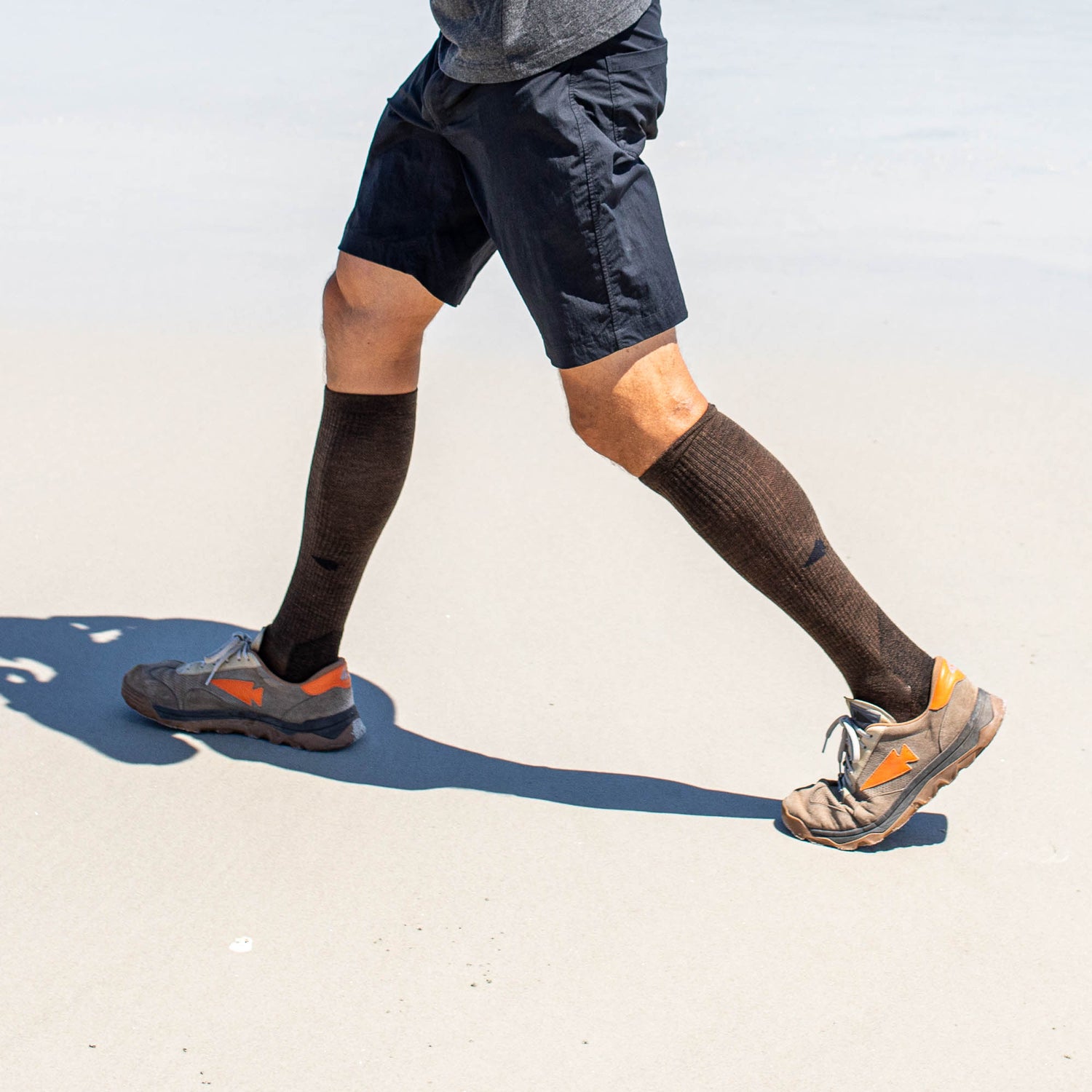 A person walks on sand wearing Merino Compression Socks in brown, paired with shorts and sneakers; upper body is not visible.