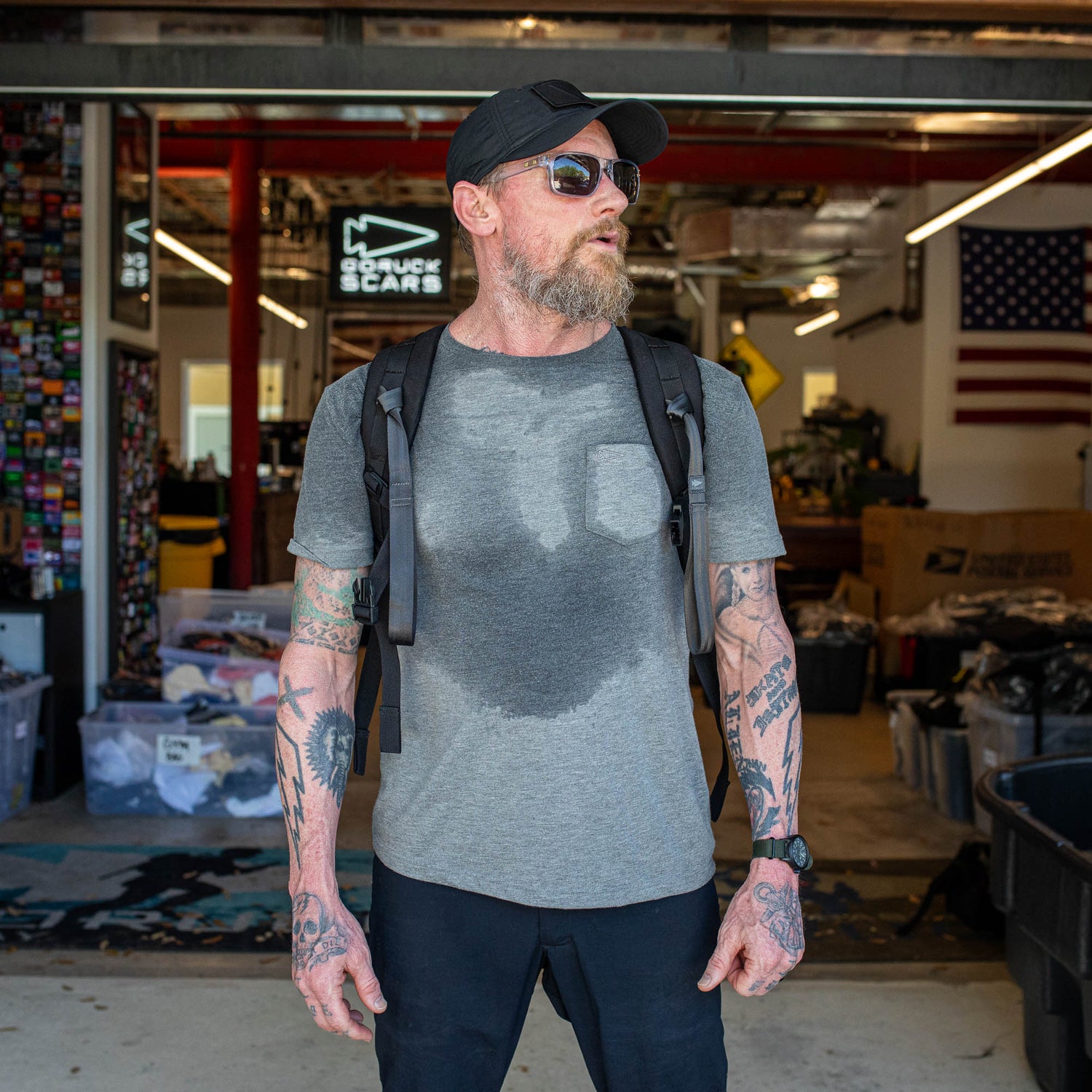 A bearded, tattooed man in sunglasses stands in a garage workspace, wearing the Short Sleeve Pocket Tee - Tri-BlendX under a backpack.