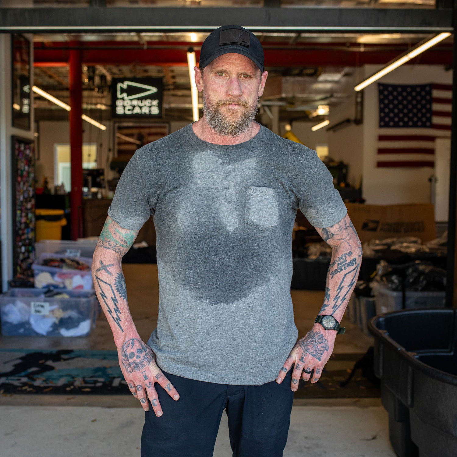 A man with tattooed arms stands indoors, his gray Short Sleeve Pocket Tee - Tri-BlendX soaked with sweat in the shape of a heart.