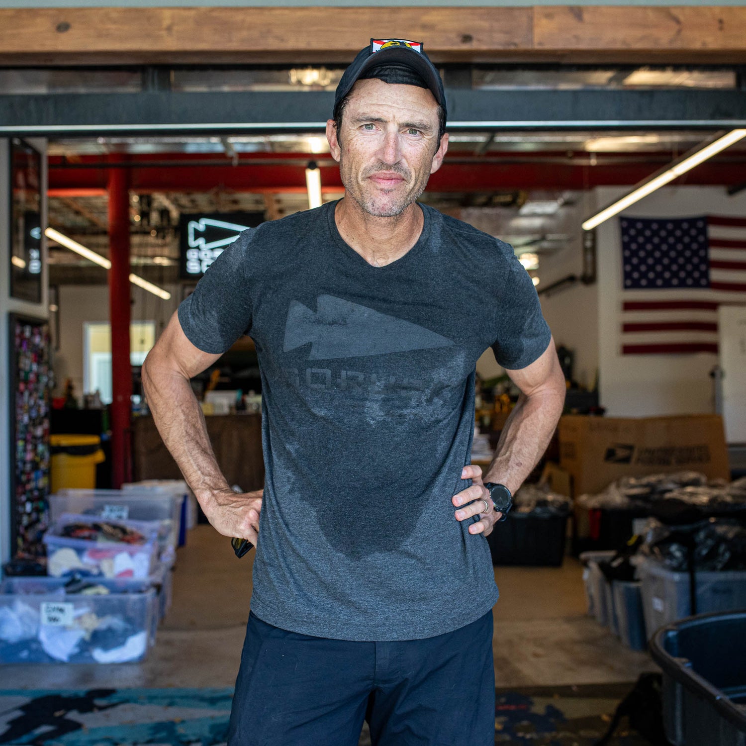 A man in a GORUCK Spearhead Tee - Tri-Blend and cap stands in a garage with storage bins and an American flag, embodying the spirit of the GORUCK spearhead.