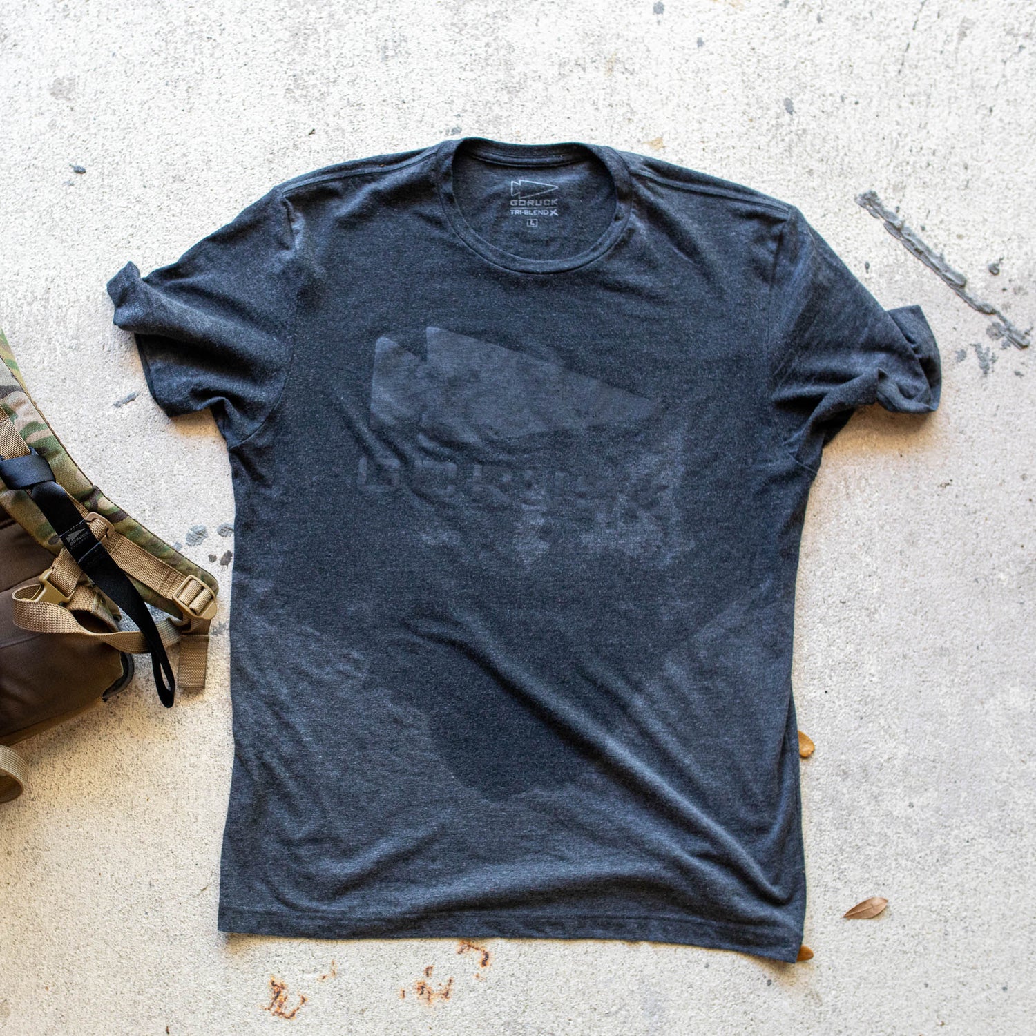The GORUCK Spearhead Tee - Tri-Blend, in dark gray, with moisture marks, rests on a concrete surface beside a camouflage bag. The shirt displays the iconic spearhead logo honoring Special Forces heritage.