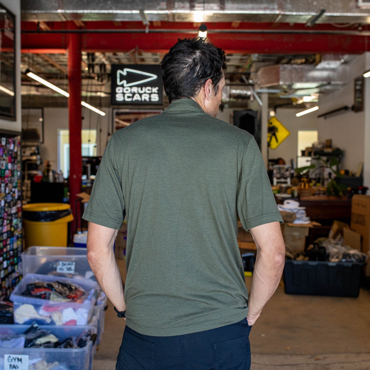 A man wearing the Men's Polo with Pocket - Tri-BlendX, made from anti-stink fabric, stands with his back to the camera in a warehouse-style workspace filled with bins and equipment.