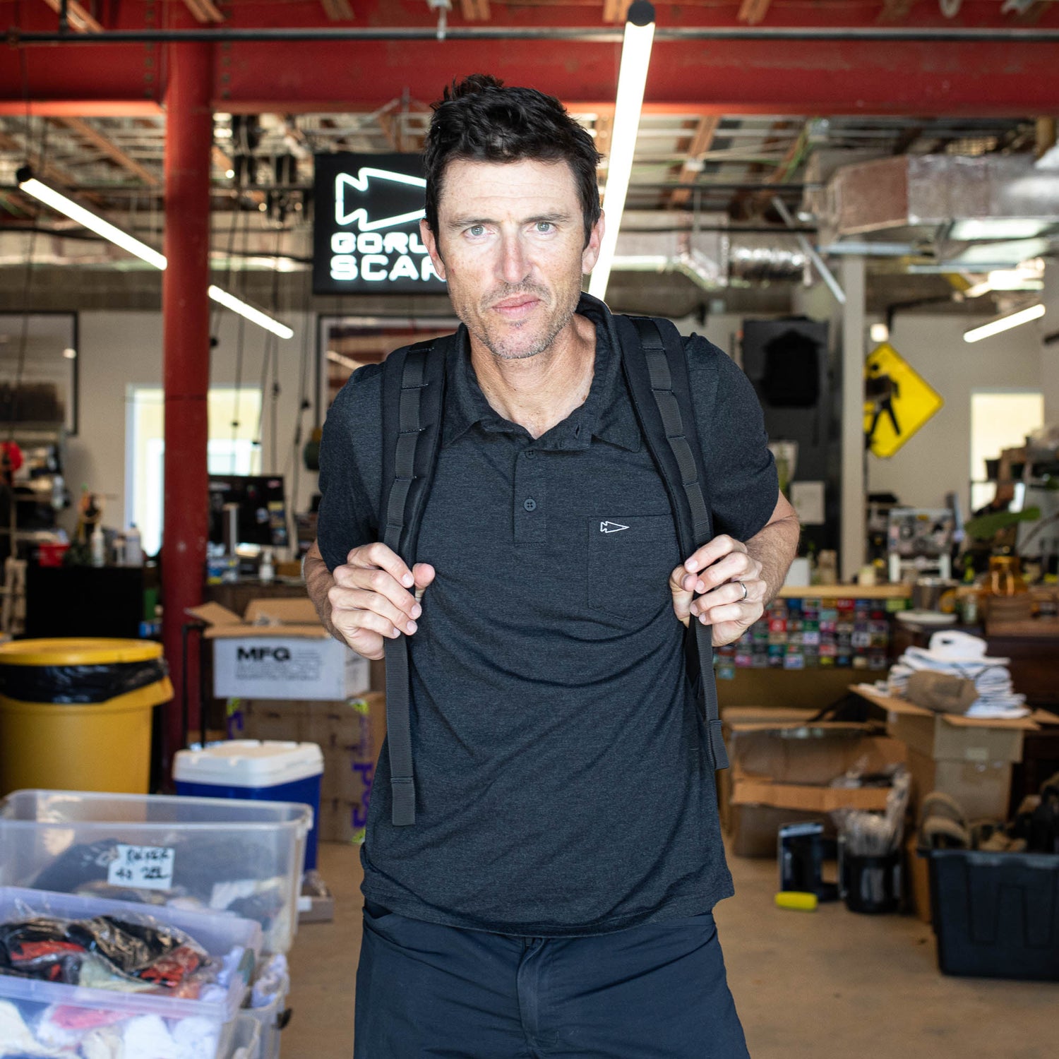 A man with a backpack stands in a cluttered office, wearing the Men's Polo with Pocket - Tri-BlendX, made from anti-stink fabric—ideal for enduring long GORUCK Events.