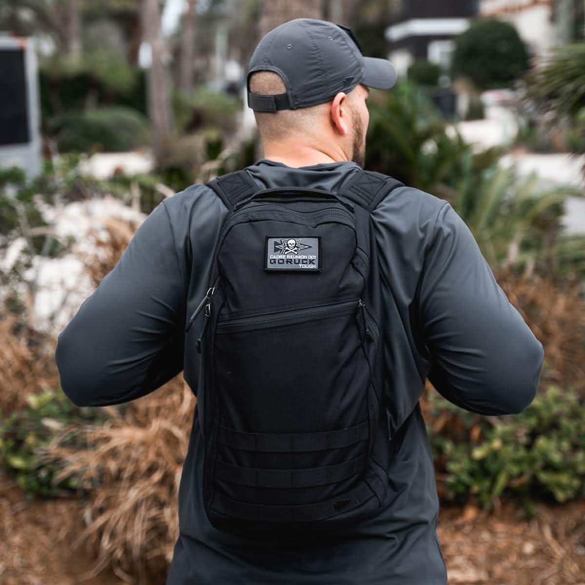 Man wearing black GORUCK backpack and dark performance shirt outdoors among greenery and urban setting