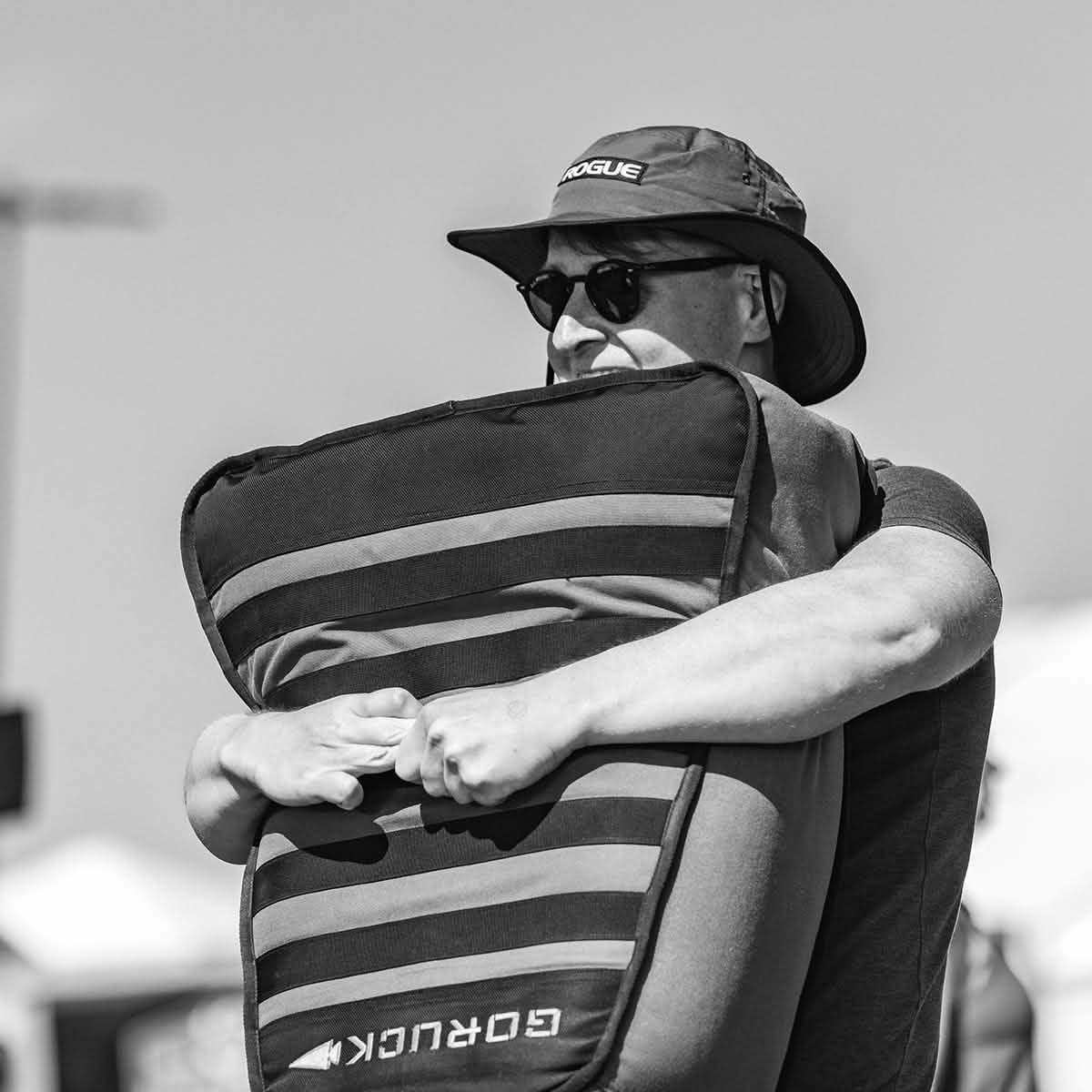 Man wearing Rogue hat and sunglasses holding a GORUCK rucking sandbag outdoors