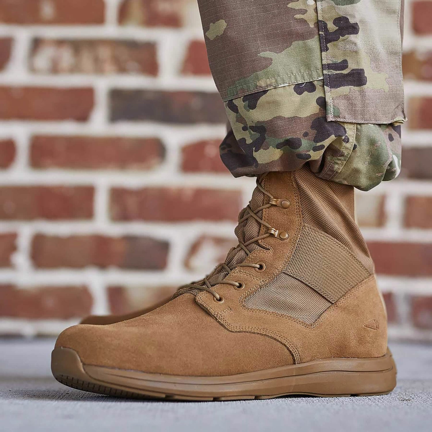 A person wearing MACV-1 Hi-Speed - High Top boots by GORUCK, which are AR670-1 compliant in tan and paired with camouflage pants, stands on a concrete surface in front of a brick wall. The lightweight and reinforced design of these high-top rucking boots is securely laced up.