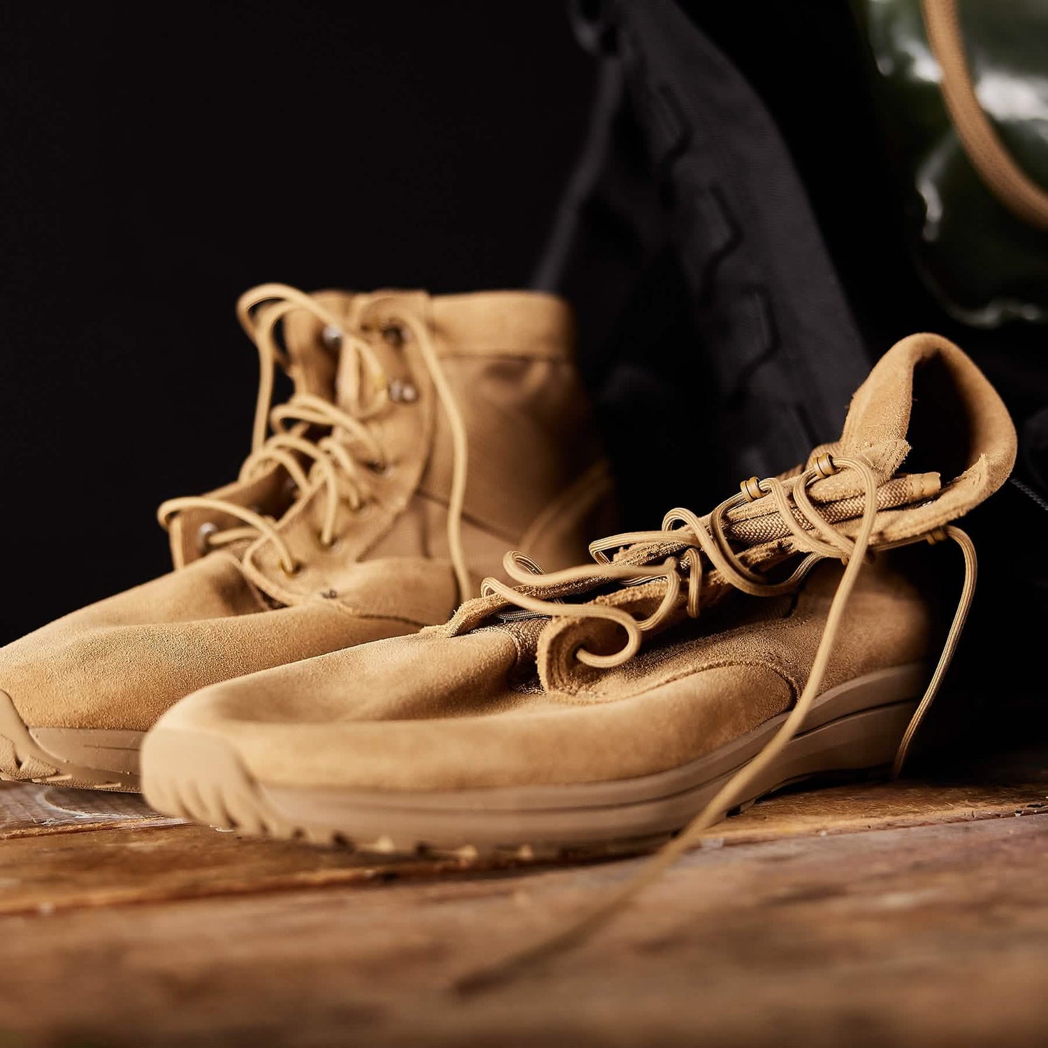 A pair of GORUCK MACV-1 Hi-Speed - Mid Top boots, featuring a lightweight design and crafted from light brown suede with laces, resting on a wooden surface. One boot is tilted to reveal the sole. A black cloth or bag is partially visible against a dark backdrop in the background.