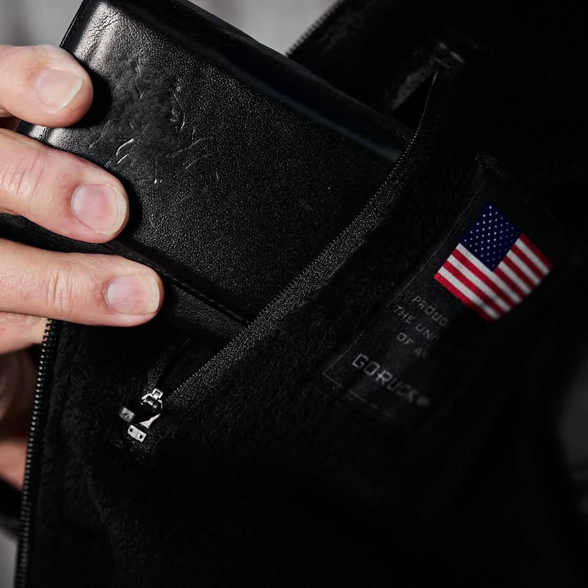 Hand placing black leather wallet inside a black GORUCK jacket's zippered chest pocket with an American flag label