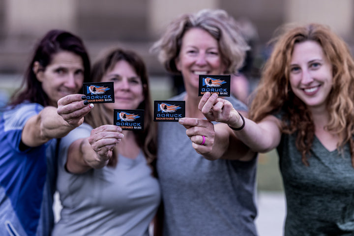 Four smiling women hold GORUCK patches toward the camera, standing together outdoors during the Badass Babes - Savannah, GA - Event Weekend 9/25-9/26 for a women-only getaway and team building event.