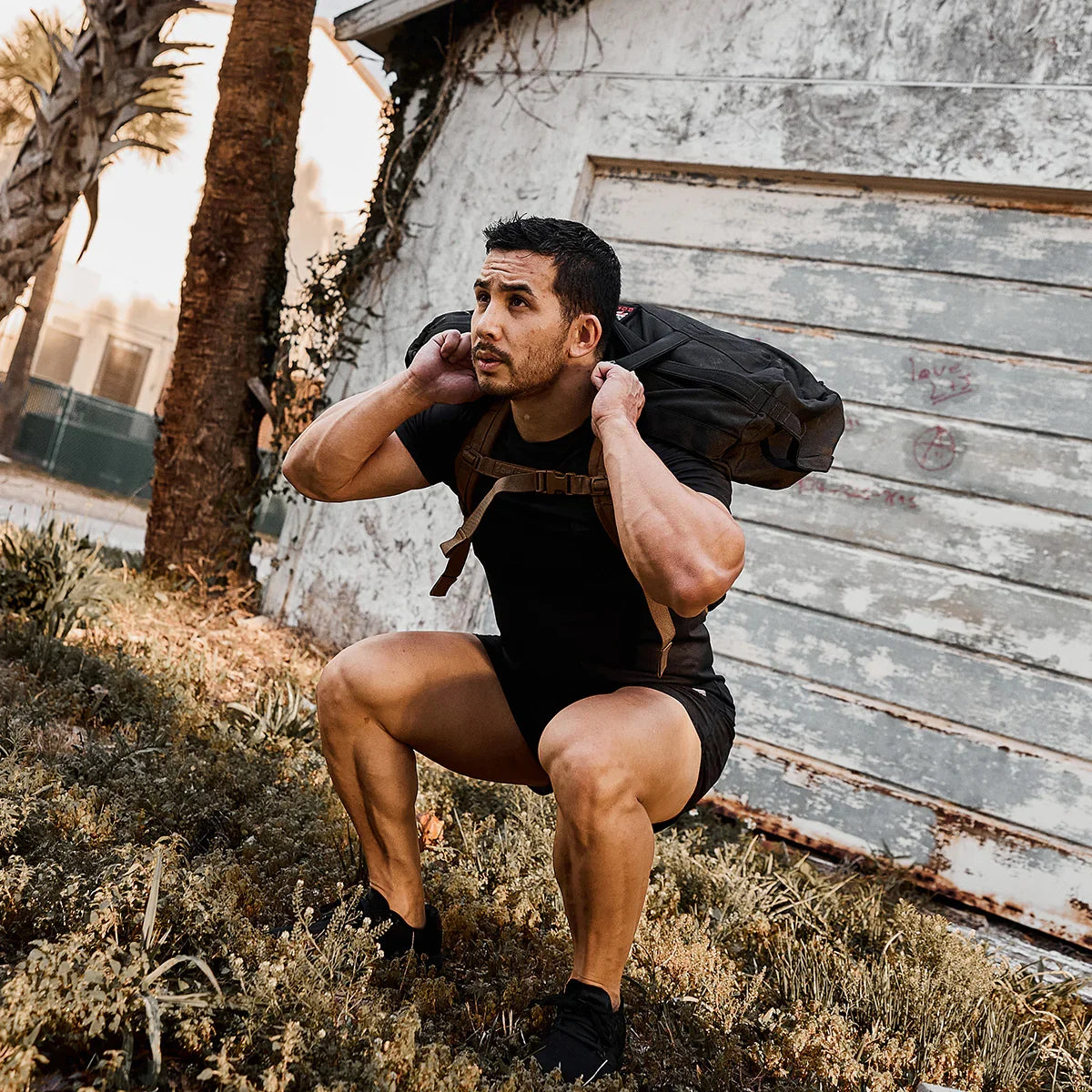 Athletic man performing squat exercise outdoors with GORUCK pack on shoulders near weathered building
