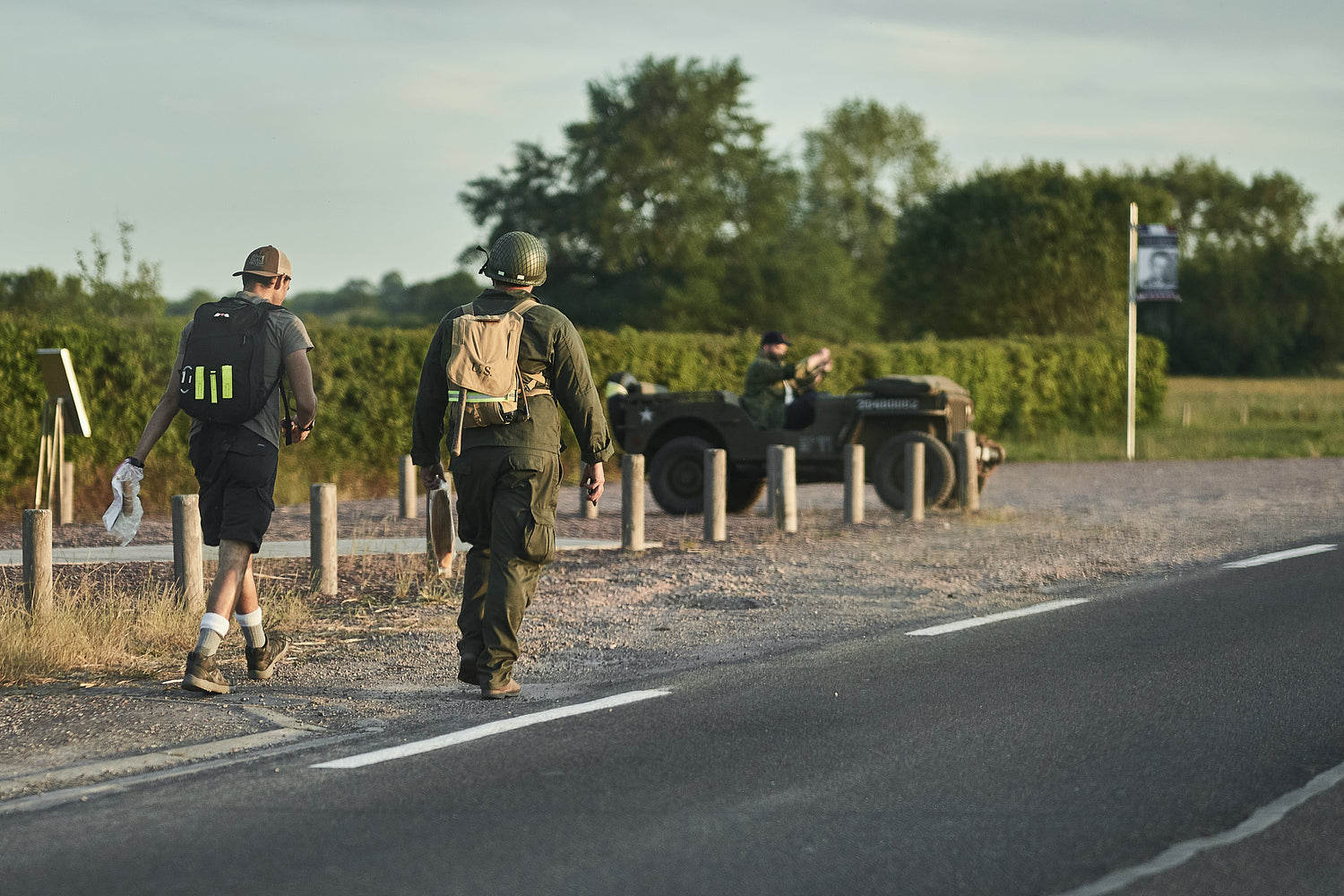 Two people with backpacks walk by a roadside; a person sits in a military jeep in the background.