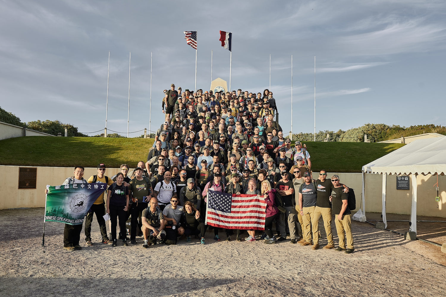 A large group poses on stairs outdoors, holding flags, under a sunny sky with two flags flying above.