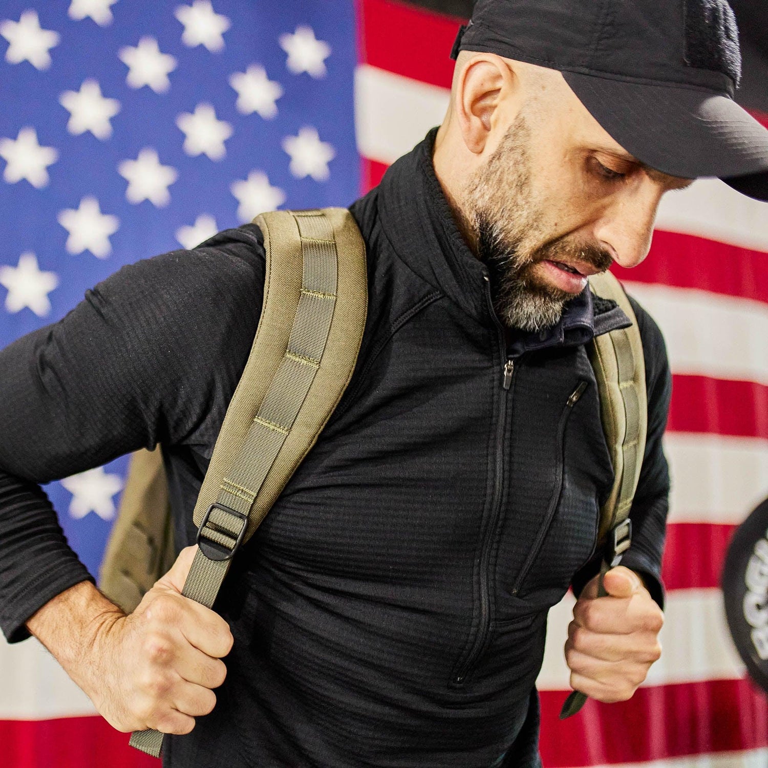Man wearing black jacket and tactical backpack straps adjusting gear with American flag backdrop, GORUCK brand