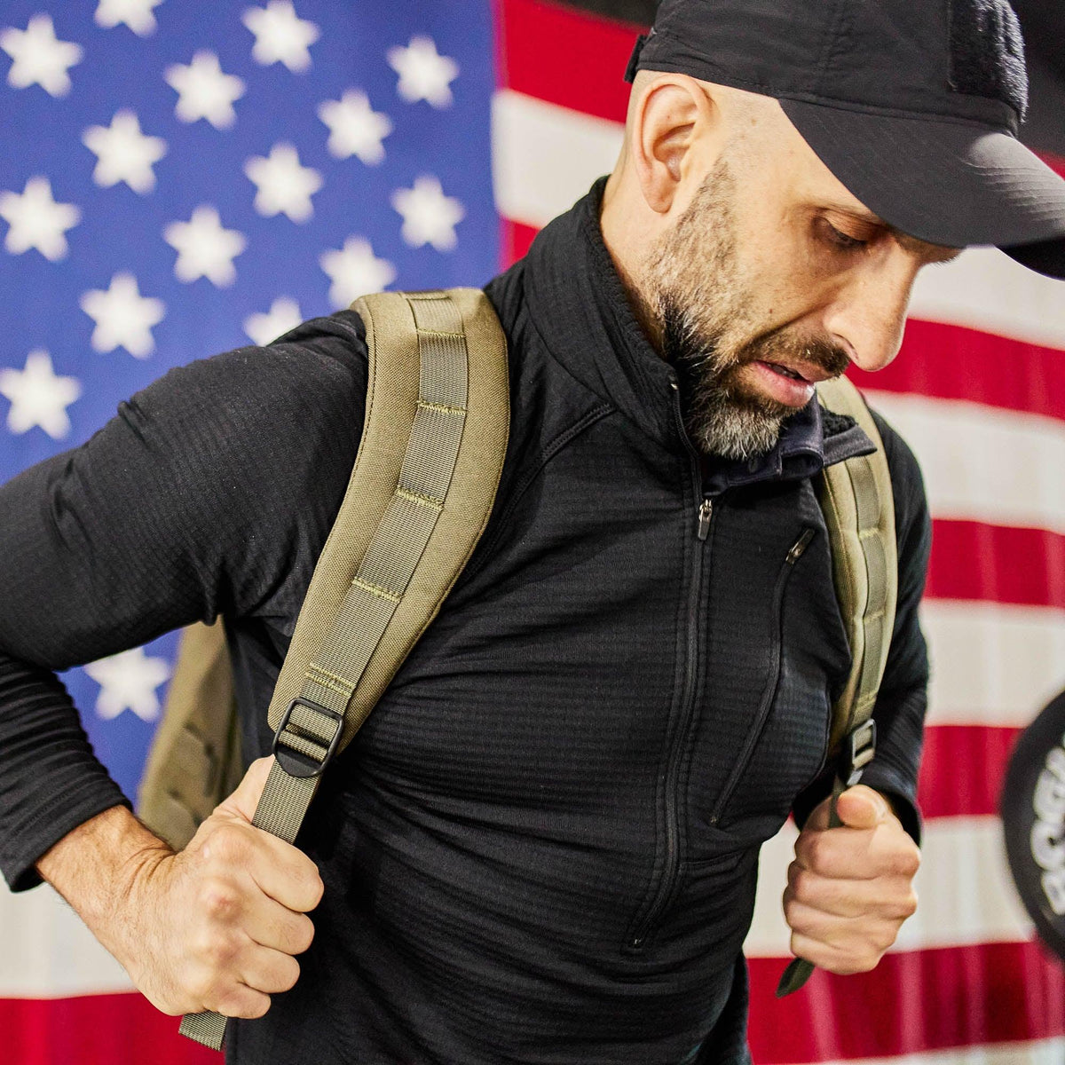 Man wearing GORUCK rucking backpack and black jacket in front of American flag