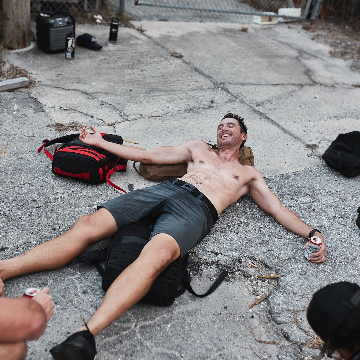 Shirtless man lying on concrete outdoors, smiling with a drink can in hand and friends nearby—his Spearhead Nylon Webbing Belt slung casually at his side, ready for the next adventure.