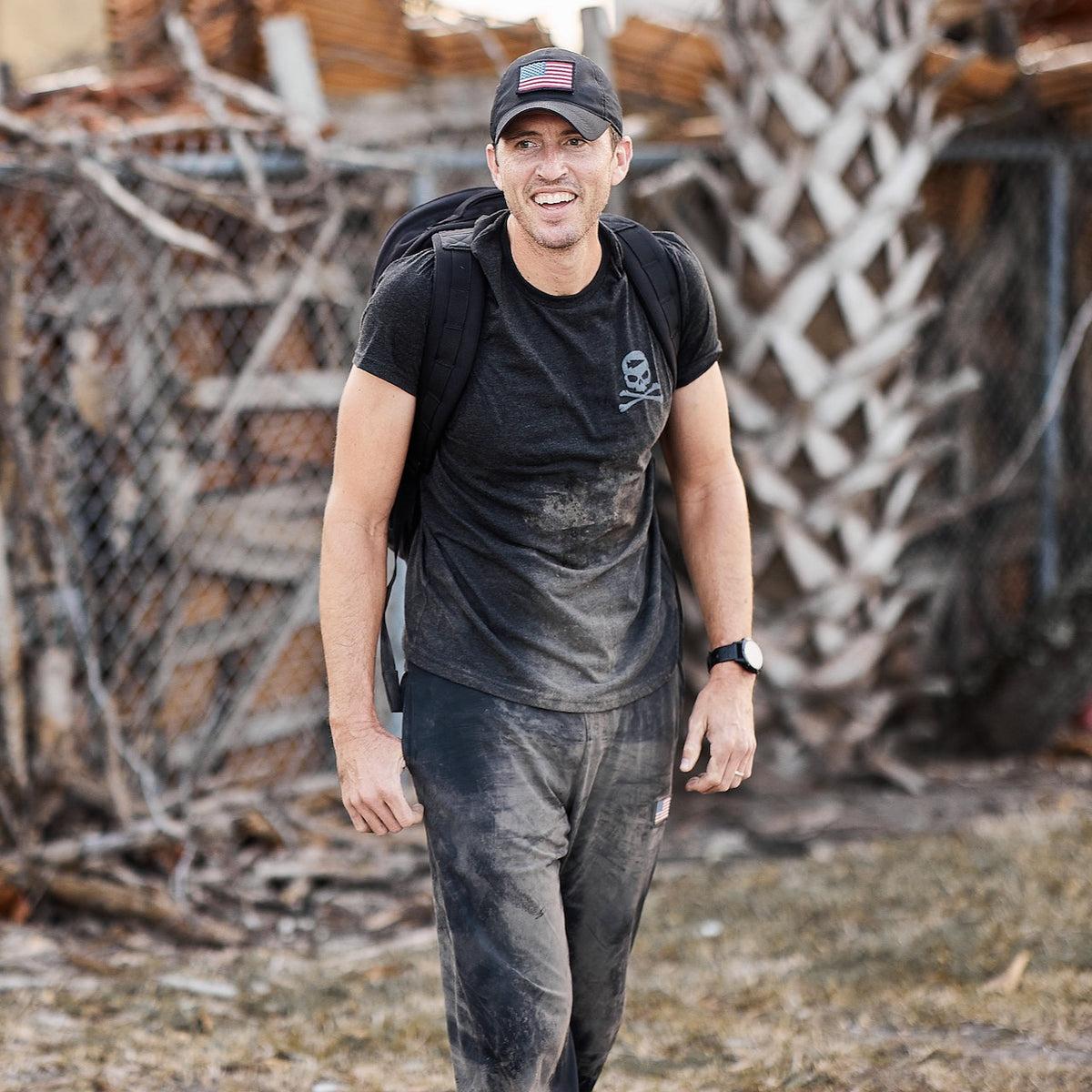 A man in a black shirt and cap stands smiling outside, wearing a backpack and the Men’s American Training Sweatpants made in the USA, with dirt on his clothes and arms.