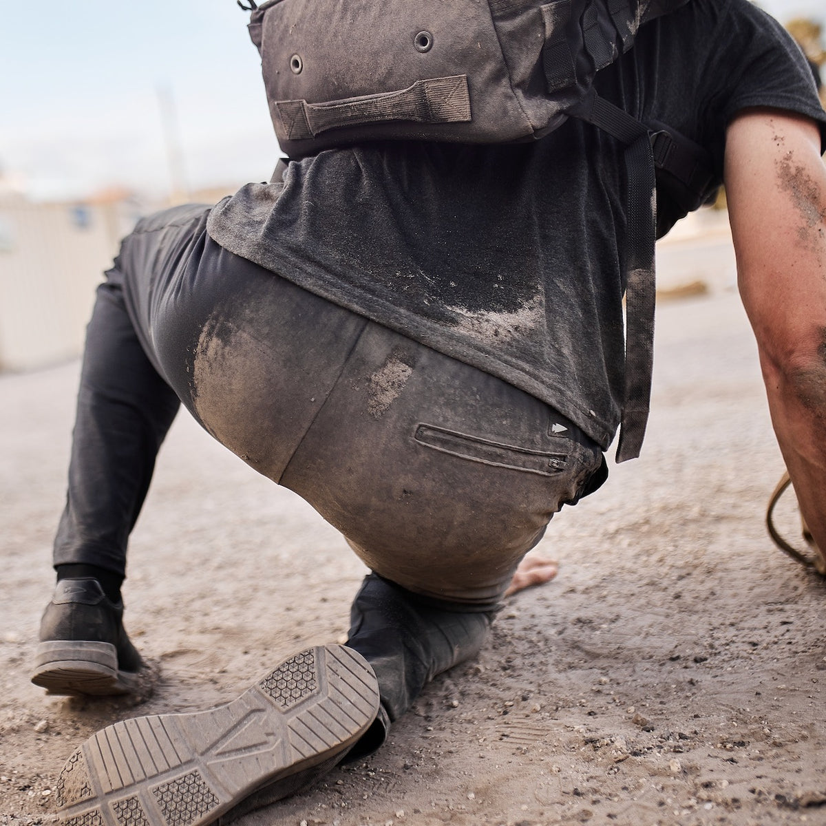 Wearing a backpack, a person kneels on gravel with dirt on their pants and hands—proof that the Men’s American Training Sweatpants are built for adventure. Made in the USA and backed by a lifetime guarantee.
