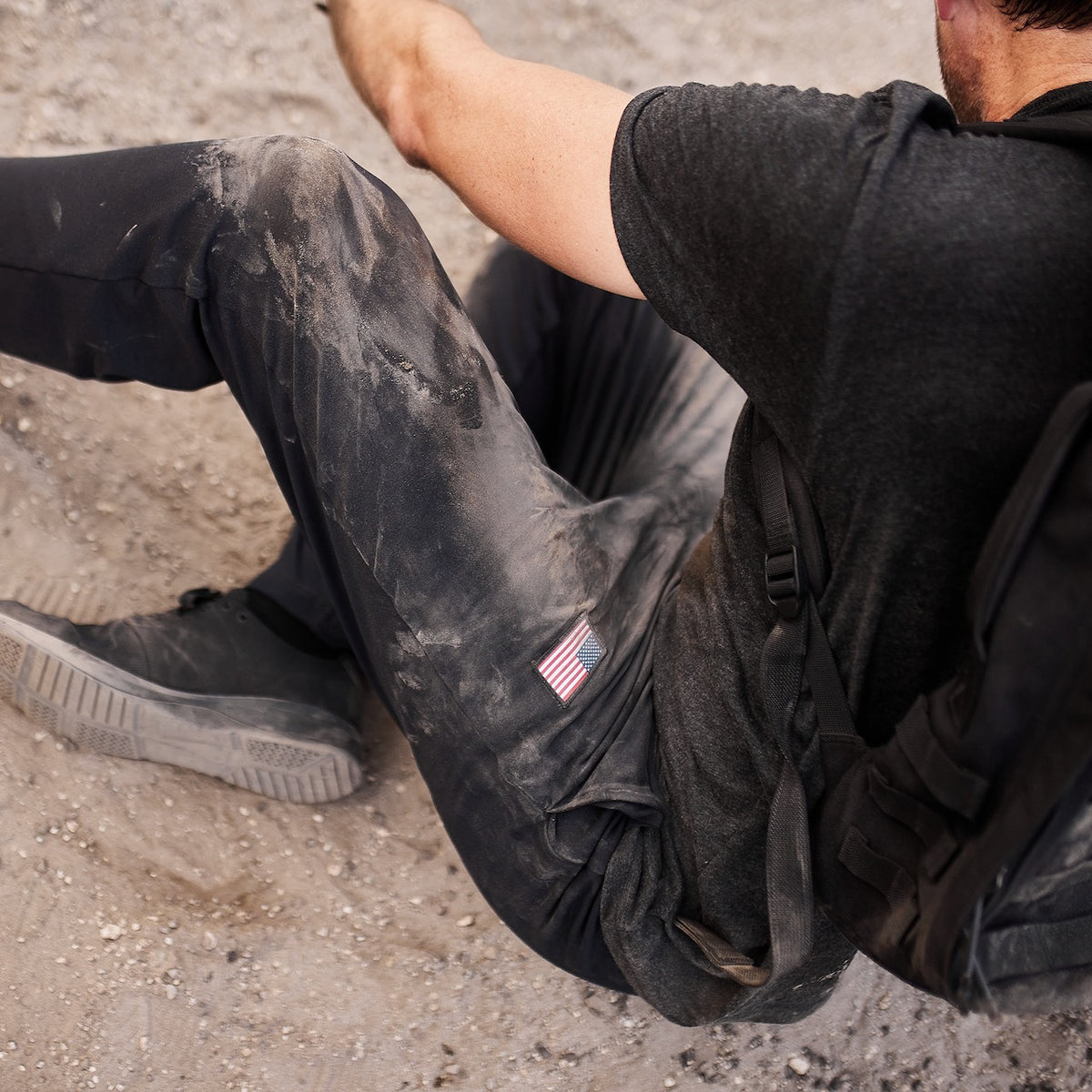 A person sits on the ground in dusty clothes, wearing Men’s American Training Sweatpants—tough, made in the USA, featuring a small US flag patch, and paired with black shoes.