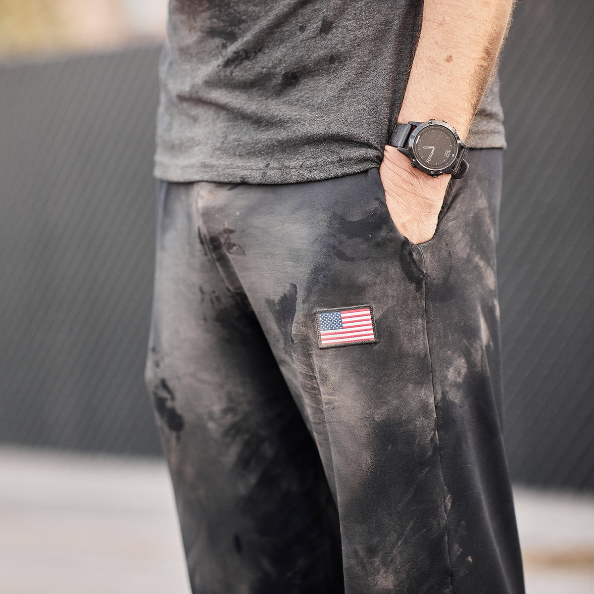 A person wearing Men’s American Training Sweatpants, featuring a tough design with an American flag patch, paired with a black watch and gray shirt visible—crafted with pride and made in the USA.