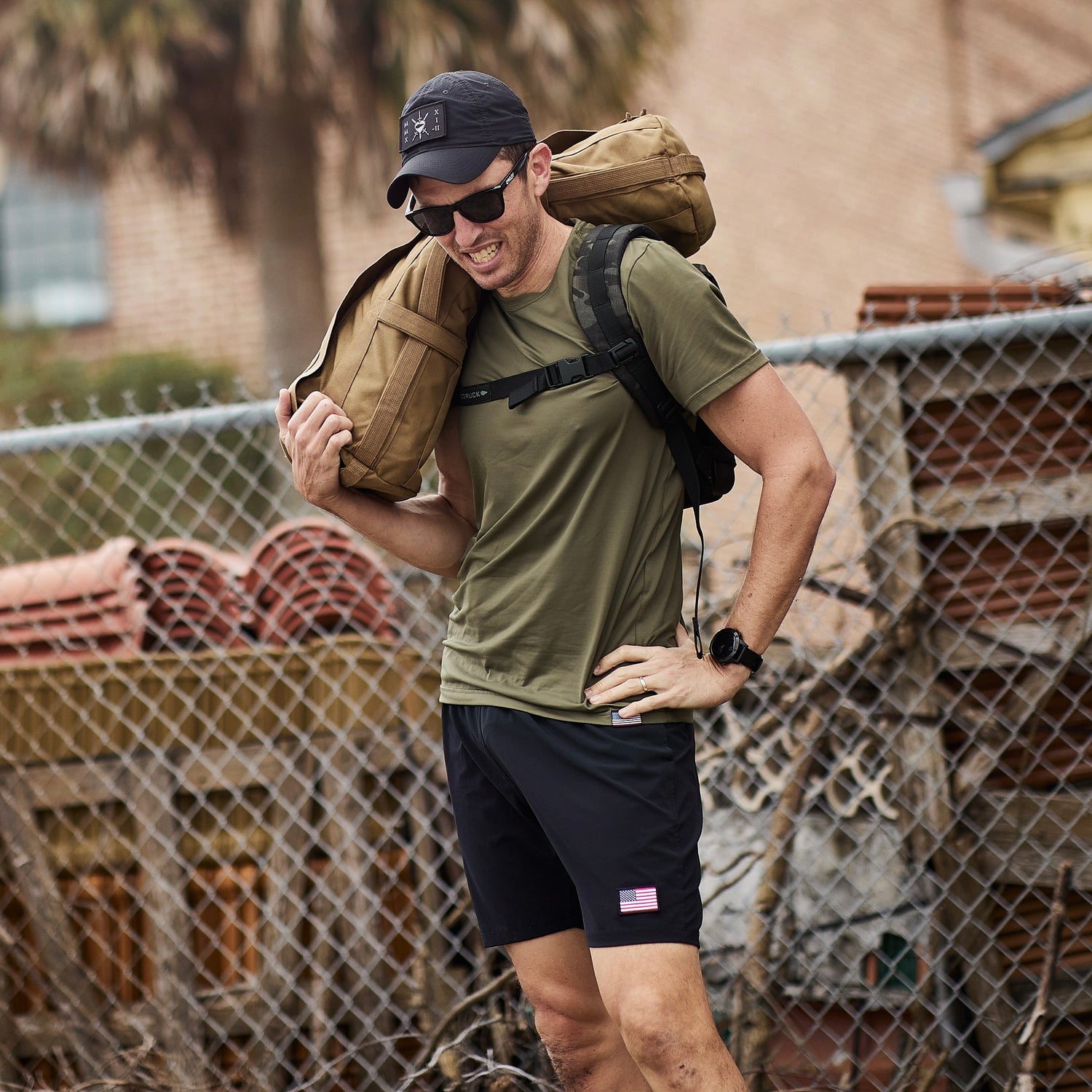 A man in athletic wear stands outdoors by a chain-link fence, carrying a sandbag over his shoulder and wearing Men’s USA Training Shorts - Flag - ToughStretch.