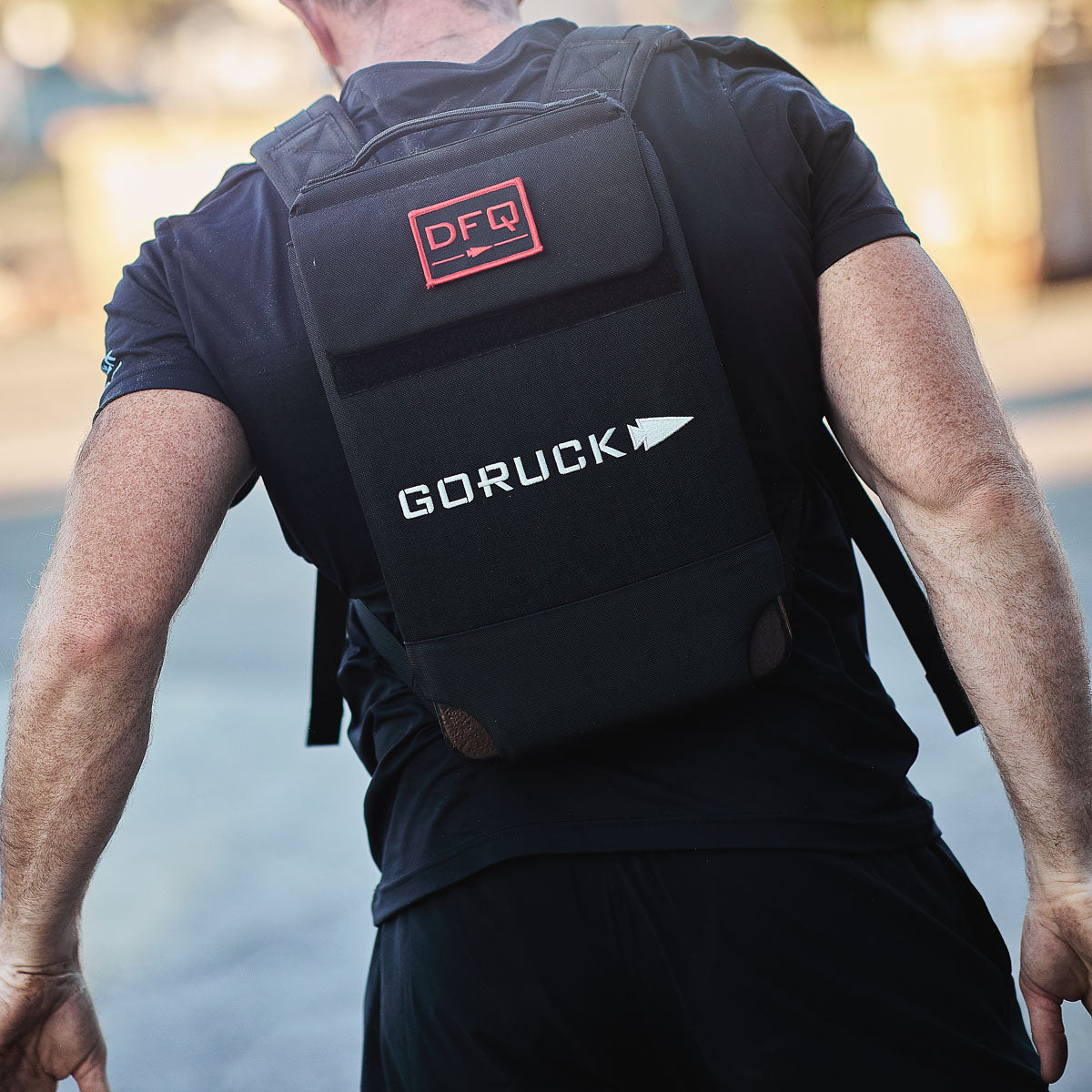 A person photographed from behind outdoors wears a black Ruck Plate Carrier 3.0 with ergonomic lumbar support and a red DFQ patch.