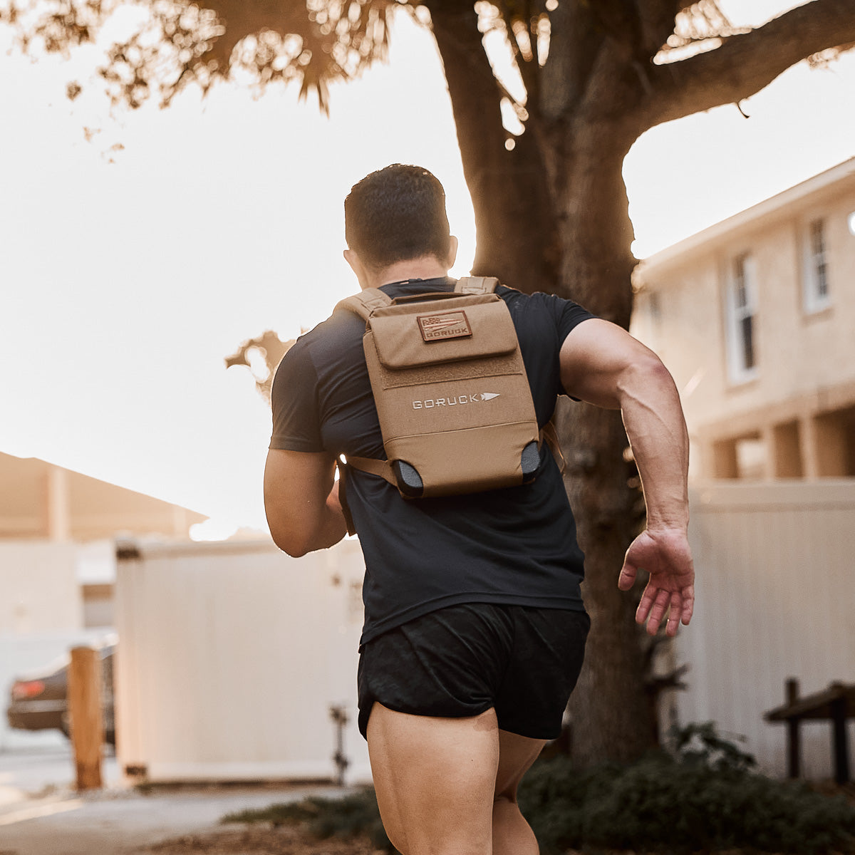A person runs outdoors wearing black shorts, a black shirt, and a tan Ruck Plate Carrier 3.0 with padded shoulder straps for extra comfort.