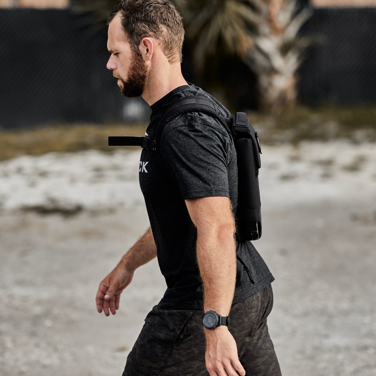 Man in workout clothes walks outdoors with a black Ruck Plate Carrier 3.0 weighted backpack featuring lumbar support and a smartwatch, against a blurred background.