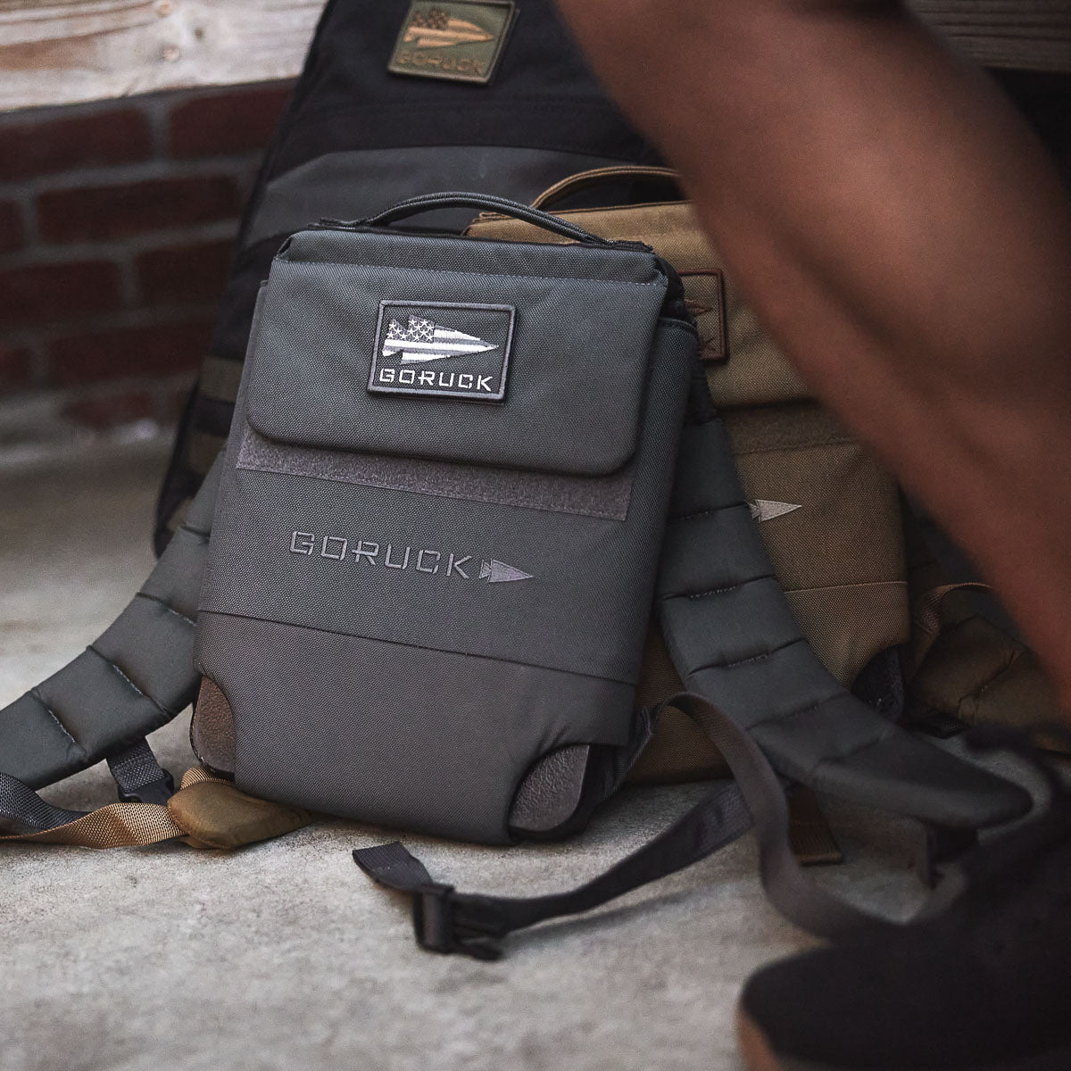 The Ruck Plate Carrier 3.0, a gray ballistic nylon backpack from GORUCK, sits on a concrete floor next to someone’s leg, resting against larger packs.