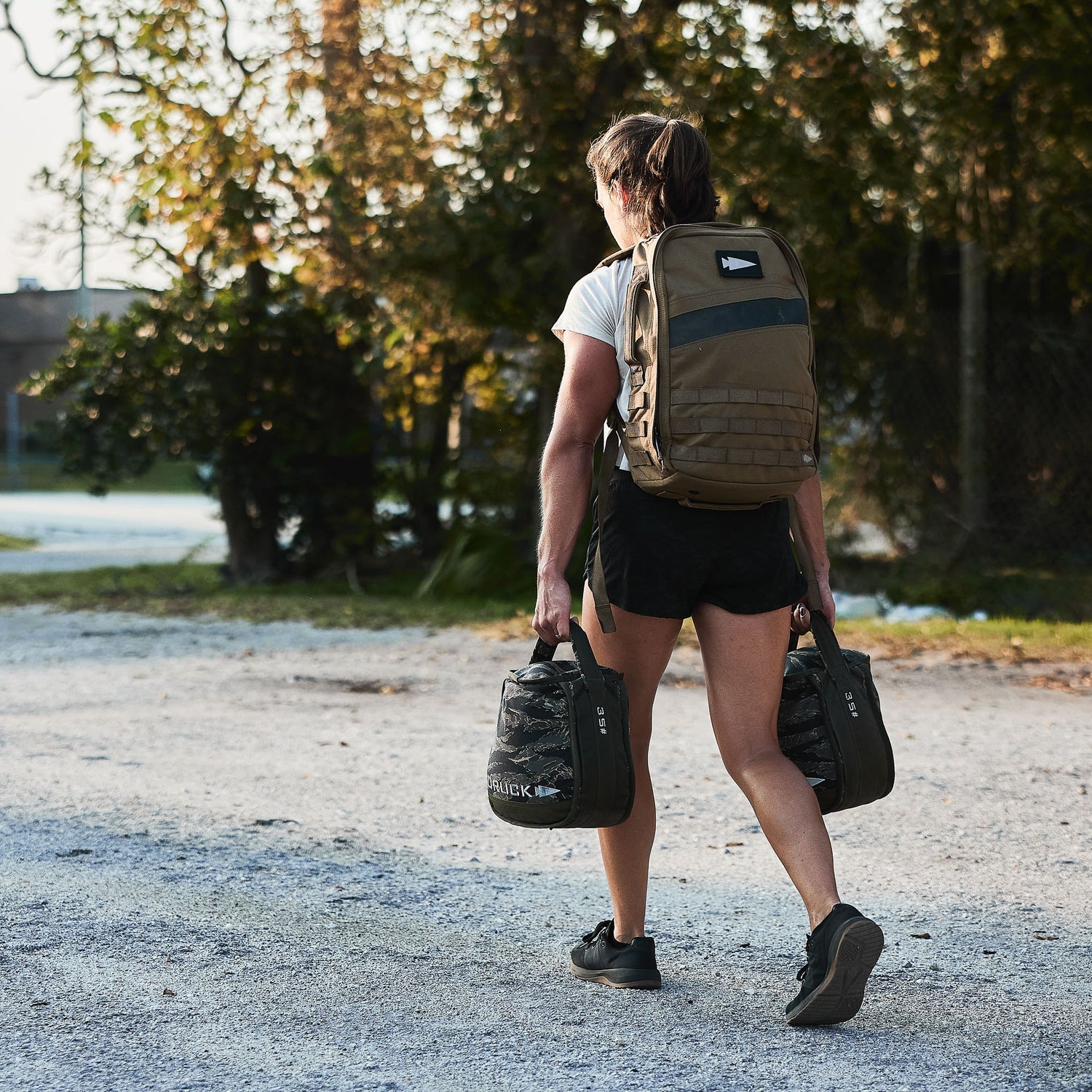 A woman with two Sand Kettlebells and a backpack walks outdoors on a gravel path, trees behind her—ideal for portable kettlebell workouts or at-home training.