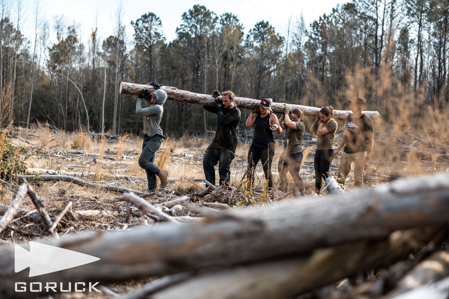 Six people carry a large log together through a wooded, clearing area with the GORUCK logo in the corner.