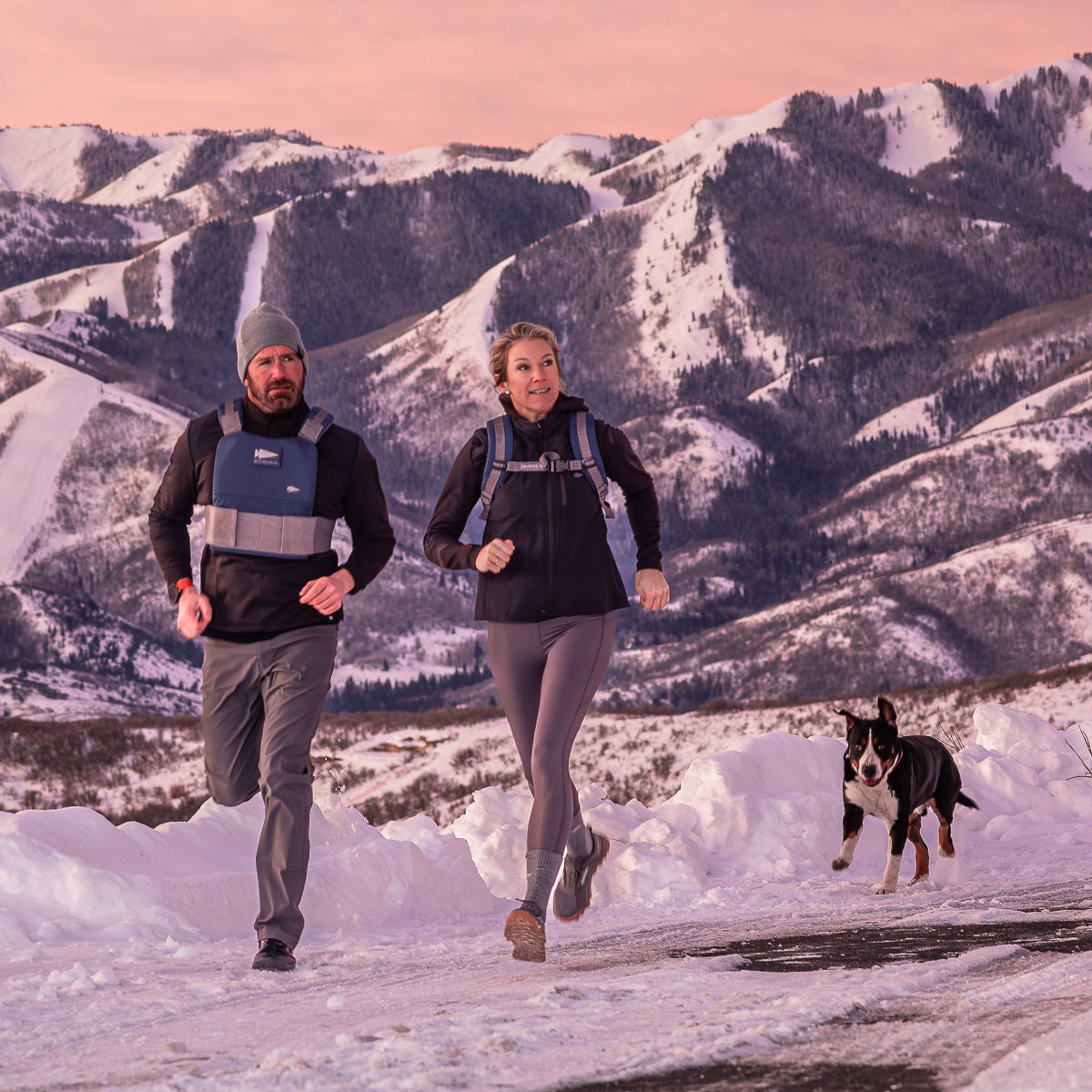 At sunset, two people and their dog run along a snowy mountain road with snow-capped peaks in the background. One runner wears the Ruck Plate Carrier 3.0, which features padded shoulder straps for extra comfort.