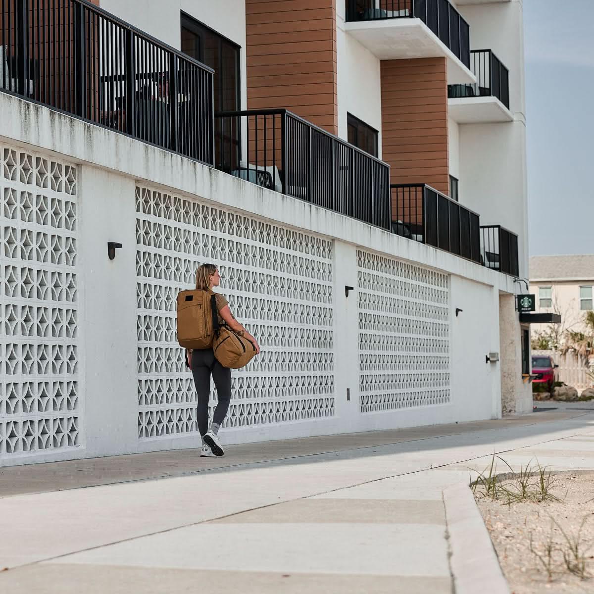 Woman carrying tan GORUCK rucksack and duffel bag walking by modern apartment building