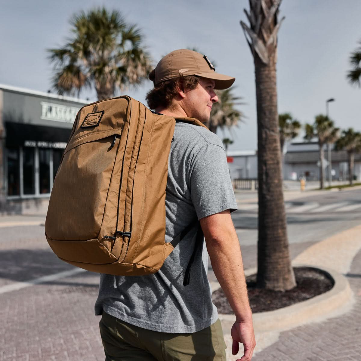 A man carrying the GR2 X-PAC backpack and wearing a cap walks outdoors near palm trees on a sunny, waterproof-perfect day.