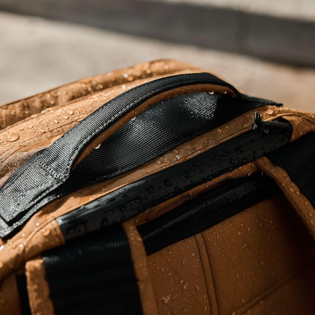 Close-up of water-resistant tan GORUCK backpack handle with black strap covered in water droplets