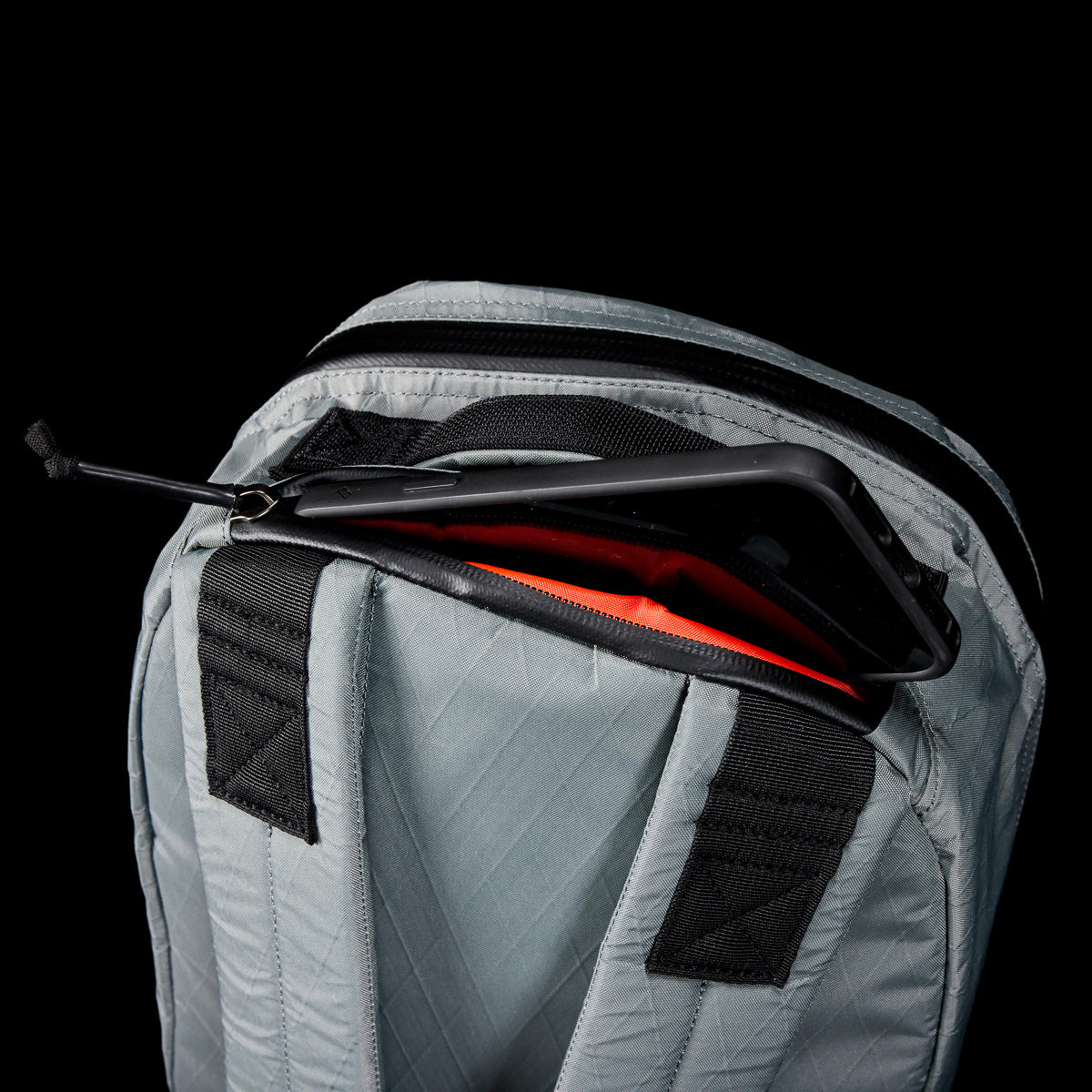 Gray Bullet X-PAC daypack with a phone partially visible in a zippered pocket, set against a black background.