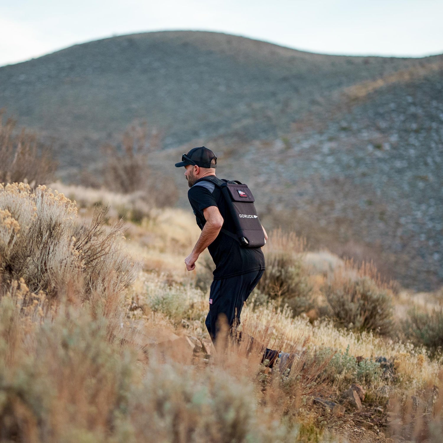A man wearing the Ruck Plate Carrier 3.0 with ergonomic lumbar support runs up a dry, grassy hillside with mountains in the background.