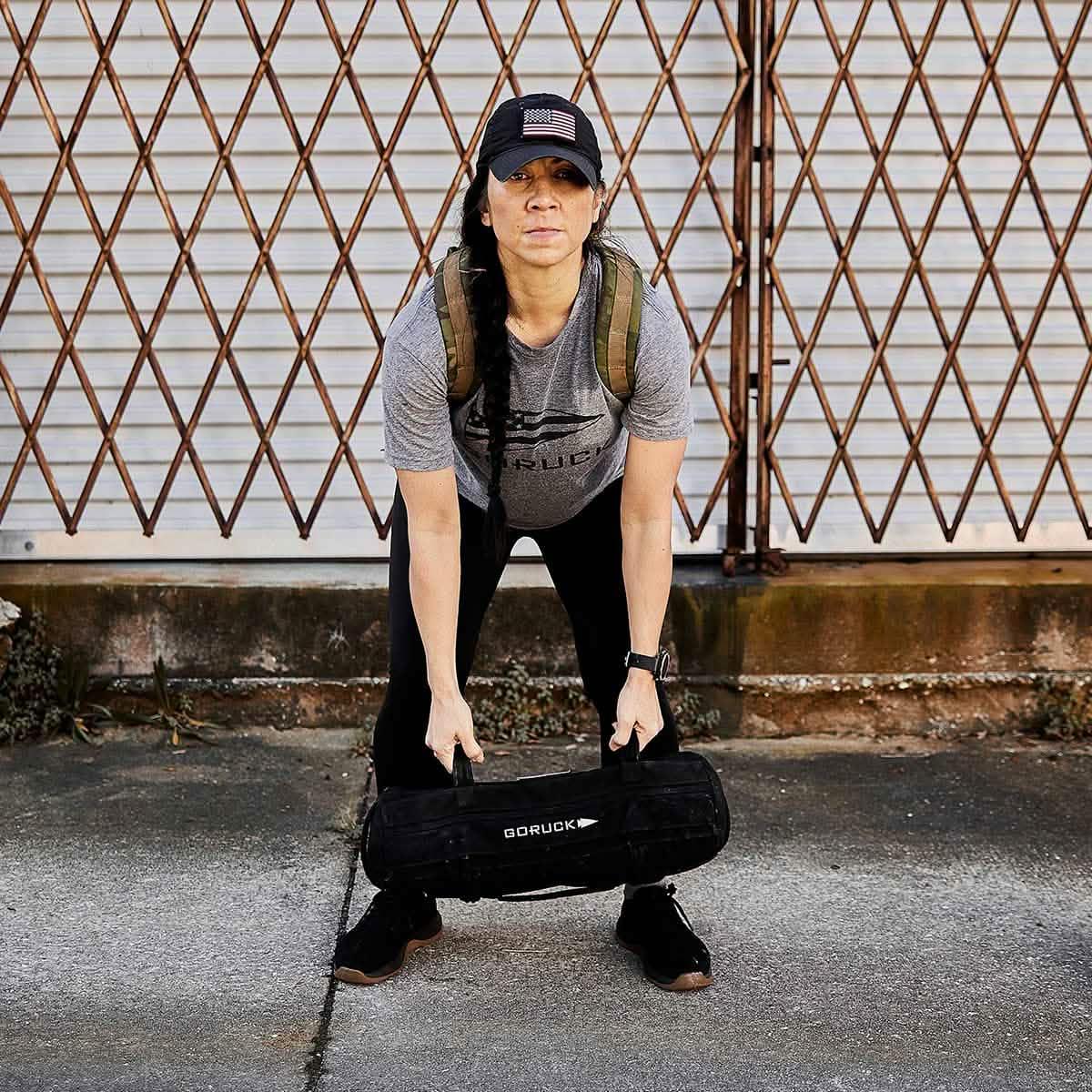 Woman wearing GORUCK gear lifting a black GORUCK sandbag outdoors against a metal lattice gate