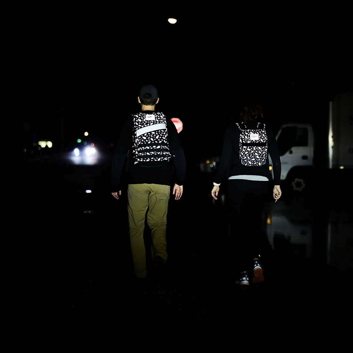 Two people with Rucker 4.0 - Reflective Spearhead rucksacks walk at night on a dark street, distant lights shining ahead.